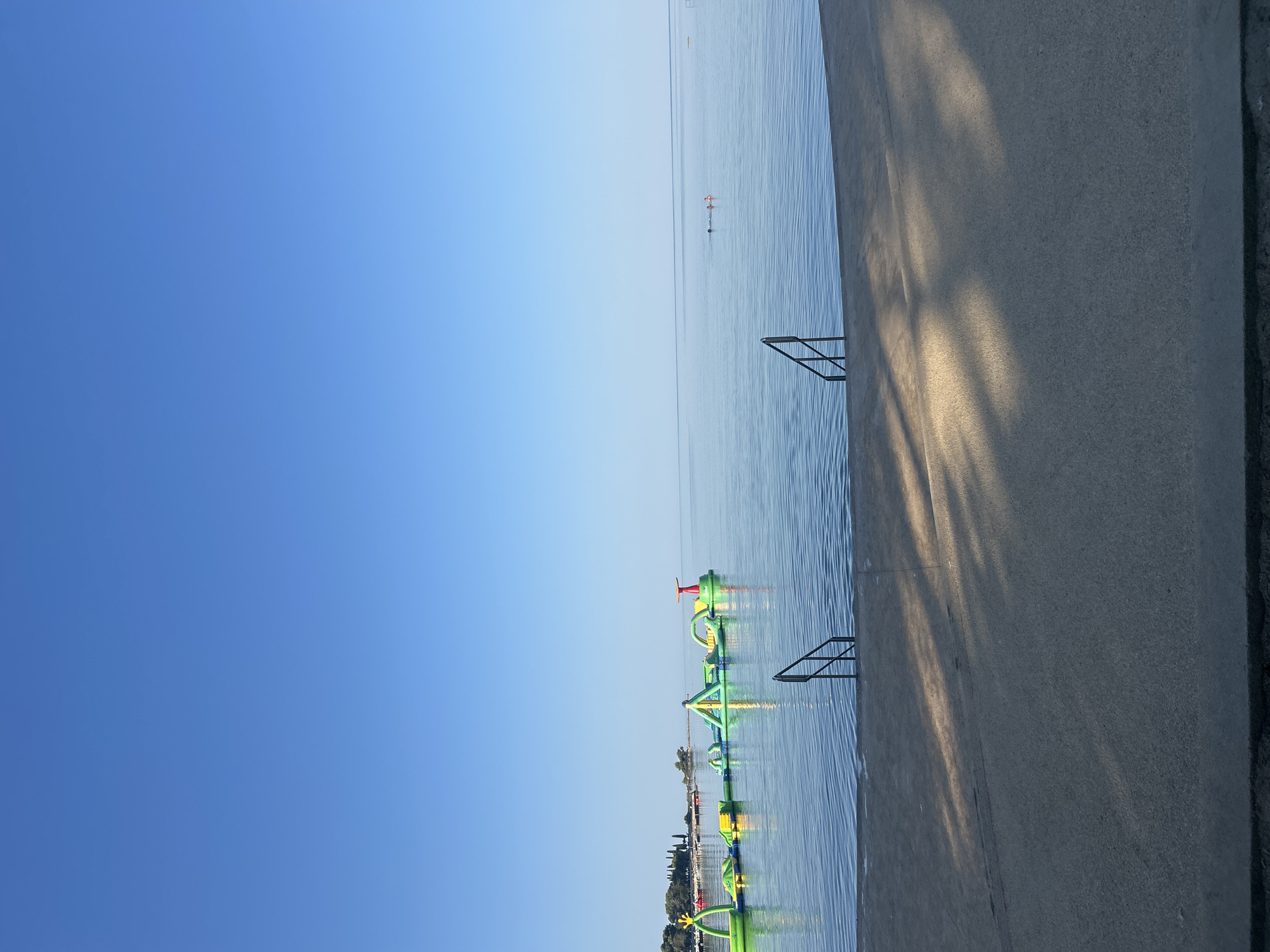 This is a landscape photograph of the sea in Katoro, Croatia. The scene depicts a bright, clear sky above a calm body of water. In the distance, a long, inflatable water park is set up in the sea, featuring various green and yellow structures, including slides and platforms. Two people are visible swimming in the water further out. In the foreground, a concrete walkway borders the water, with a set of metal stairs leading down into the sea. The walkway is in shadow in some areas, suggesting the presence of overhead trees or structures casting dappled light. The overall impression is one of a peaceful, sunny day at a seaside resort.