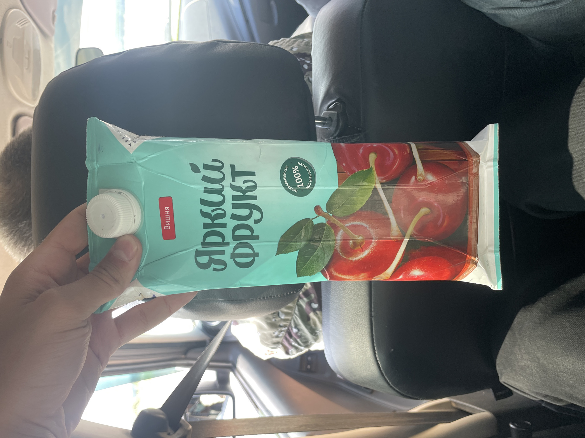 A person's hand holds a teal-colored juice carton with a white cap. The carton features a picture of ripe cherries and Russian text that reads "ЯРКИЙ ФРУКТ" (Bright Fruit) and "ВИШНЯ" (Cherry). A red label on the carton states "НАТУРАЛЬНЫЙ СОК 100% (Natural Juice 100%)". The background shows the interior of a car with black leather seats, a headrest, and a window with a view of greenery outside. The scene appears to be a casual moment, likely during a car ride. The location is not determinable from the image.