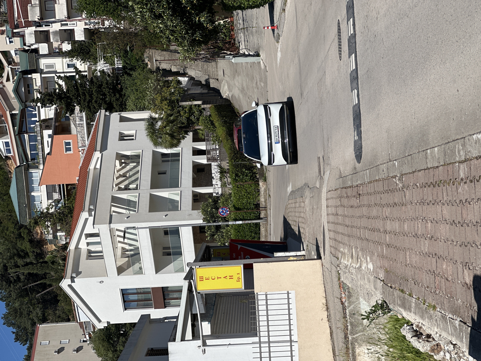 The image captures a sunlit street scene in a residential area of Budva, Montenegro. A white car, likely an SUV or crossover, is visible on the asphalt road, facing away from the viewer. Its Bosnian license plate, "BH 9079 YA", is clearly discernible. The road features a black and white striped speed bump and a metal drain cover. To the right of the road, a brick-patterned sidewalk runs alongside a low wall.
Multi-story residential buildings with various architectural styles line both sides of the street. Many of these structures are light-colored, featuring multiple balconies with glass or striped railings. Some buildings have terracotta-colored roofs, typical of the region. Lush green trees and dense bushes are abundant, interspersed among the buildings and along the roadside, casting distinct shadows under the bright sunlight.
In the foreground on the left, a prominent yellow rectangular sign displays Cyrillic text that reads "ШЕСТАН Бр.1". Below and to the right of this sign, a red and black sign is partially visible, showing fragments of text including "Get ATM" and "OMAT". A circular road sign with a red border, blue background, and white symbols stands near these signs. No people are visible, and the scene appears quiet and still under clear, sunny weather conditions.