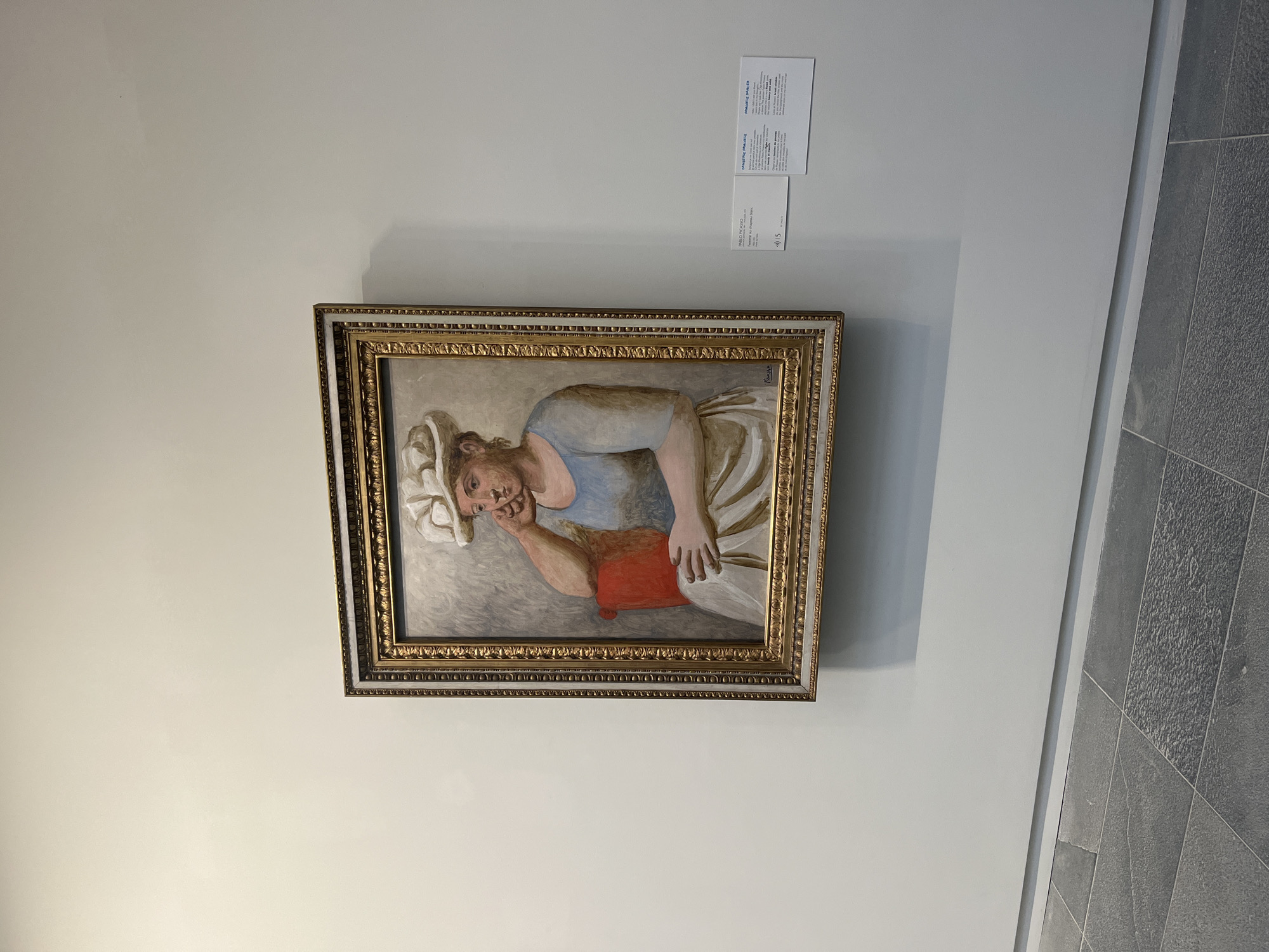 A framed painting is prominently displayed on a plain, light-colored wall in what appears to be an art gallery or museum. The painting, oriented in portrait style, depicts a woman in a reclining or resting pose. She is wearing a light blue top and a white or cream-colored skirt or lower garment. A white cloth or turban is wrapped around her head. Her head is gently supported by her right hand, which rests on a reddish-orange cushion or pillow. The woman has an intense, perhaps weary or contemplative expression, with her eyes open and looking slightly upwards. The painting has a muted color palette, with earthy tones for the skin and background, contrasting with the blue, white, and the pop of red.
The artwork is encased in an ornate, gold-colored frame with intricate decorative patterns. To the upper right of the painting, two white informational cards are taped to the wall. The text on these cards, largely in French, identifies the artist as PABLO PICASSO. One card clearly states "LAVEUSE IMPERIALE" (Imperial Washerwoman) and "La Laveuse, 1917," followed by "Femme au Chapeau Blanc" (Woman in a White Hat). The other card repeats "Femme au Chapeau blanc" and has "0/15" at the bottom right.
The setting appears to be an indoor exhibition space, possibly in Paris, France, given the French text. To the right of the painting, a gray tiled floor is visible, further reinforcing the museum or gallery environment. The lighting suggests an indoor setting, possibly with overhead artificial light, creating subtle shadows on the wall.