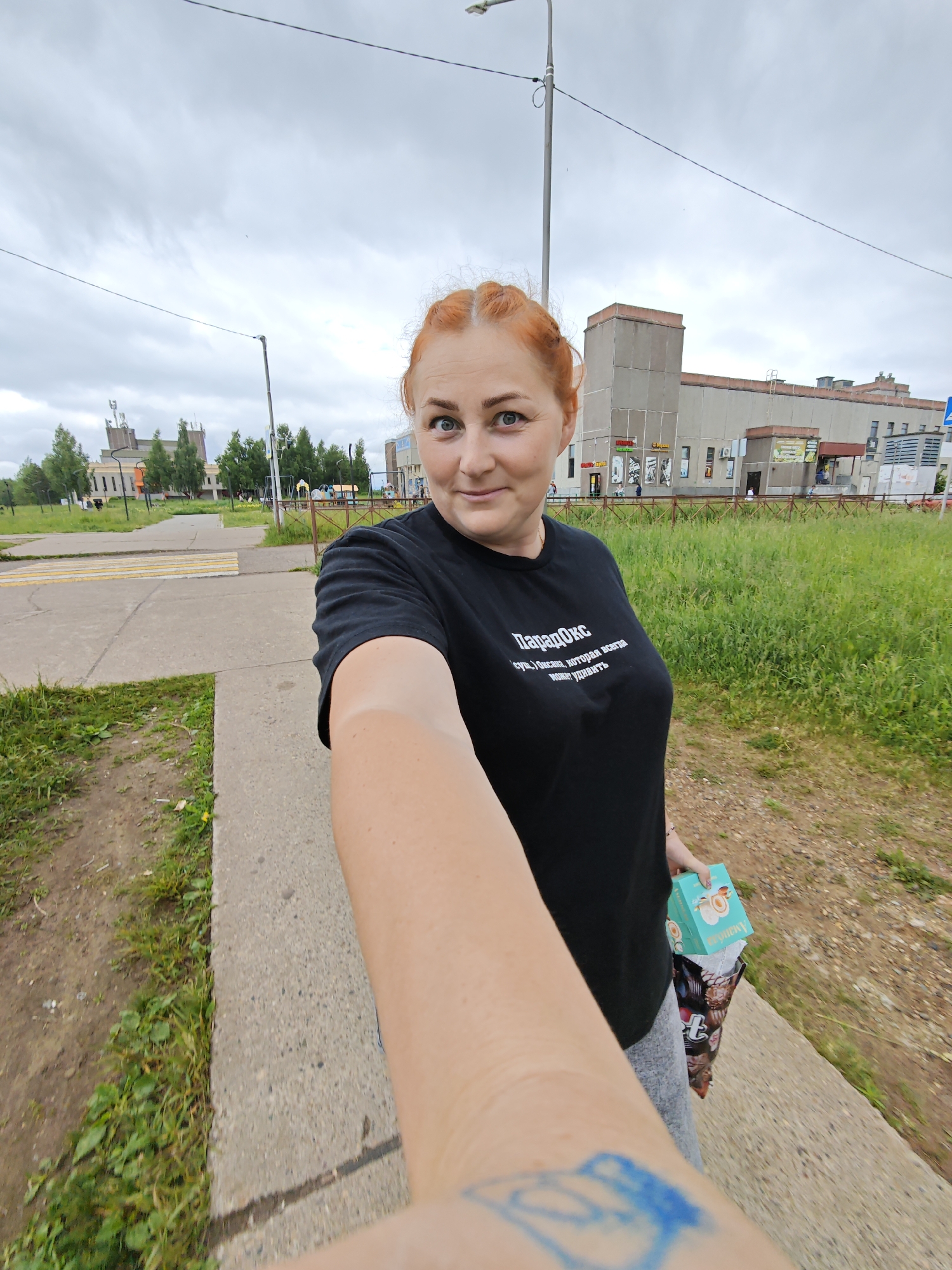 A red-haired woman poses for a selfie on an overcast day in Марковский, Russia. She wears a black T-shirt with the Russian word "ПарадОкс" (Paradox) and a definition below it, "то, что всегда может удивить" (that which can always surprise), along with light gray bottoms. Her right arm, extended to hold the camera, shows a prominent tan line and a partially visible blue tattoo on her forearm. She has a subtle smile/smirk and blue eyes. In her left hand, she carries a light blue box labeled "Акварель" with a hazelnut icon, and a colorful plastic bag.
She stands on a concrete sidewalk, with green grass and dirt patches on either side. A pedestrian crosswalk with yellow stripes is visible further down the path to her left. Beyond the grass, a rusted brown metal fence runs parallel to the path. In the midground, several green trees and a light pole are visible. Further back, a large, multi-story building with a flat roof and visible windows/storefronts occupies the right side of the frame. Another building with a distinctive reddish-brown, boxy structure is seen in the left midground. The sky is largely overcast with gray clouds, and electrical wires traverse the upper portion of the frame. A blue road sign is partially visible on the far right.