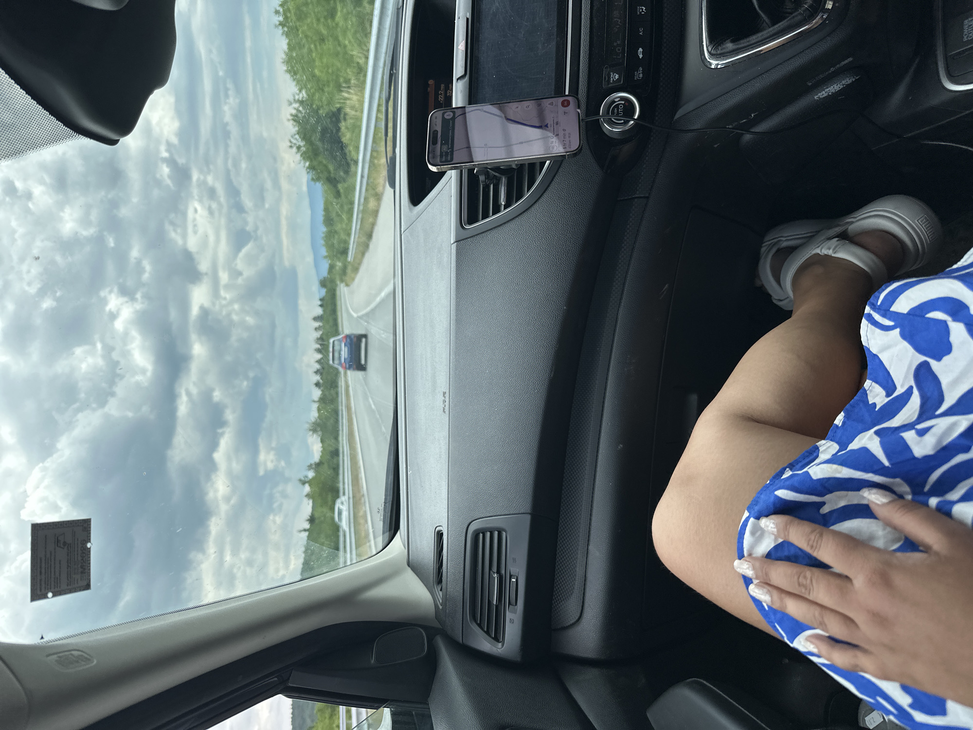 The image provides a passenger's perspective from inside a car traveling on a highway during daytime. The sky is largely overcast with scattered clouds.
In the foreground to the right, a person's lower body is visible. They are wearing a blue and white patterned outfit and white PUMA-branded sandals on their feet. Their left leg is casually propped up, and their hand, adorned with white, patterned manicured nails, rests on their lap.
The car's dark-colored dashboard occupies the middle ground. A smartphone is mounted horizontally on one of the central air vents, displaying a navigation application. The screen shows the time "22:20", a remaining distance of "222.0km", and a speed of "10.0km/h". Some Cyrillic text is visible on the map interface. A charging cable is connected to the phone. Below the phone, various car controls and buttons are visible, including "A/C", "FRONT", "AUTO", and a "POWER OUTLET 12V 180W".
Beyond the car's interior, a multi-lane highway stretches into the distance, flanked by green trees and what appear to be mountains in the background. A white guardrail runs alongside the road. Another car, dark blue in color, is visible ahead on the highway. A small, rectangular sticker, likely a vehicle registration or vignette, is affixed to the upper left side of the windshield, though its text is illegible. The scene depicts a relaxed journey through a natural landscape near Djerman, Bulgaria.
