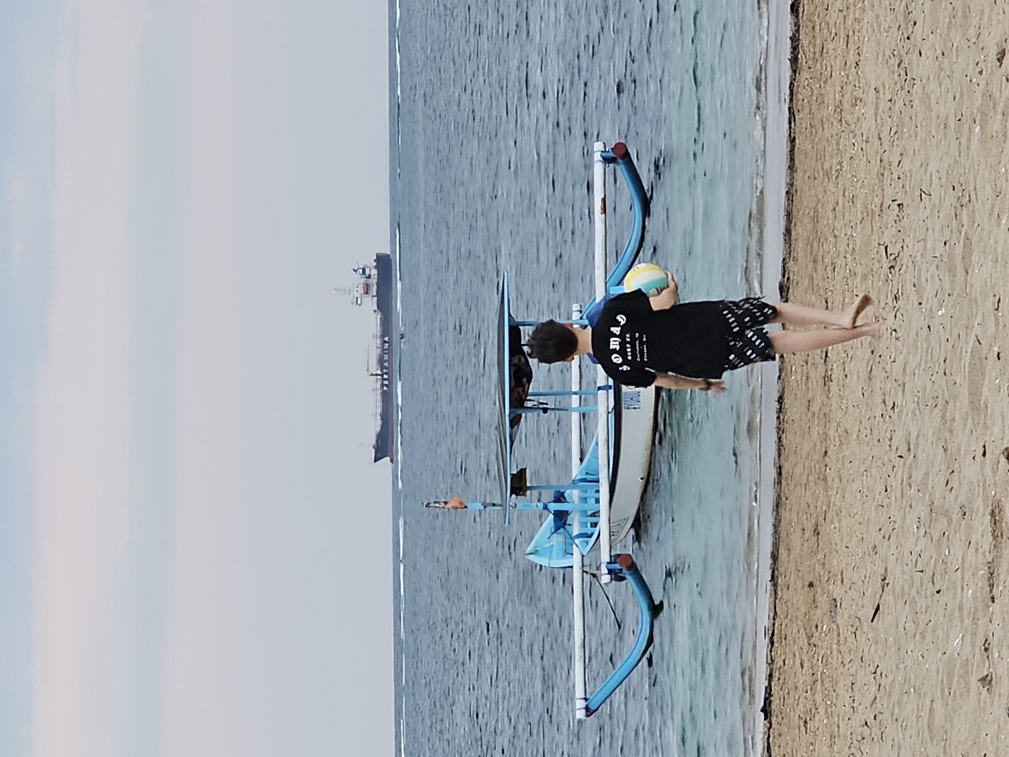 The image shows a boy in a black t-shirt and patterned shorts holding a colorful ball, walking on a sandy beach toward the ocean. In the foreground, the beach is covered with sand and small debris. The ocean is calm with small waves lapping the shore. A traditional Balinese fishing boat with blue outriggers is anchored in the shallow water. Further out in the ocean, a large cargo ship with "PERTAMINA" visible on its side is sailing. The sky is overcast with a pale blue and grey hue. The scene is likely Denpasar, Indonesia, given the characteristic outrigger boats and the presence of the Pertamina ship, which is an Indonesian state-owned oil and gas company.