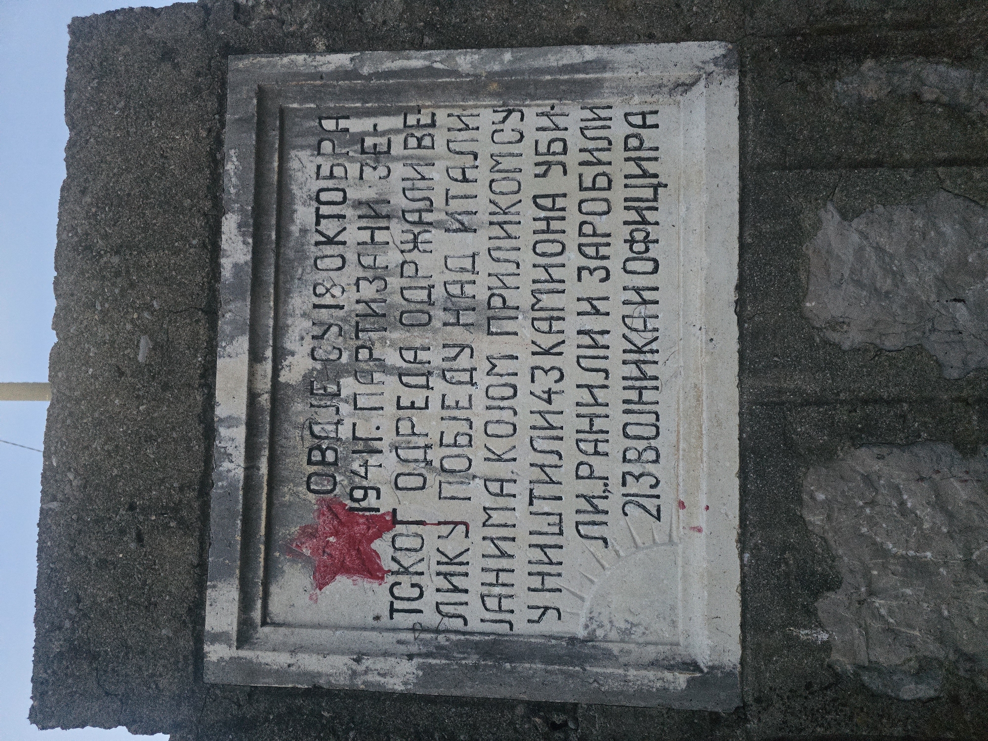 The image shows a stone memorial with Cyrillic text and a red star. The memorial is mounted on a rough concrete structure. The text is written in black, with some parts appearing faded or smudged. The red star is painted on the upper left corner of the memorial.
The text on the memorial reads:
"ОВДЈЕ СУ 18 ОКТОБРА
1941Г. ПАРТИЗANI 3E-
ТСКОГ ОДРЕДА ОДРЖАЛИ ВЕ-
ЛИКУ ПОБЈЕДУ НАД ИТАЛИ-
ЈАНИМА. КОЈОМ ПРИЛИКОМ СУ
УНИШТИЛИ 43 КАМИОНА УБИ-
ЛИ,РАНИЛИ И ЗАРОБИЛИ
213 ВОЈНИКА И ОФИЦИРА"
This translates to:
"HERE ON OCTOBER 18,
1941, THE PARTISANS OF THE
ZE-TSKOD ODRED HELD A GREAT
VICTORY OVER THE ITALIANS. DURING WHICH THEY
DESTROYED 43 TRUCKS, KILLED,
WOUNDED AND CAPTURED
213 SOLDIERS AND OFFICERS"
The location is identified as Klopot, Montenegro. The environment appears to be outdoors, possibly on a hillside, given the rough concrete and stone construction. The sky is visible in the background, appearing overcast or hazy. There are no people visible in the image.