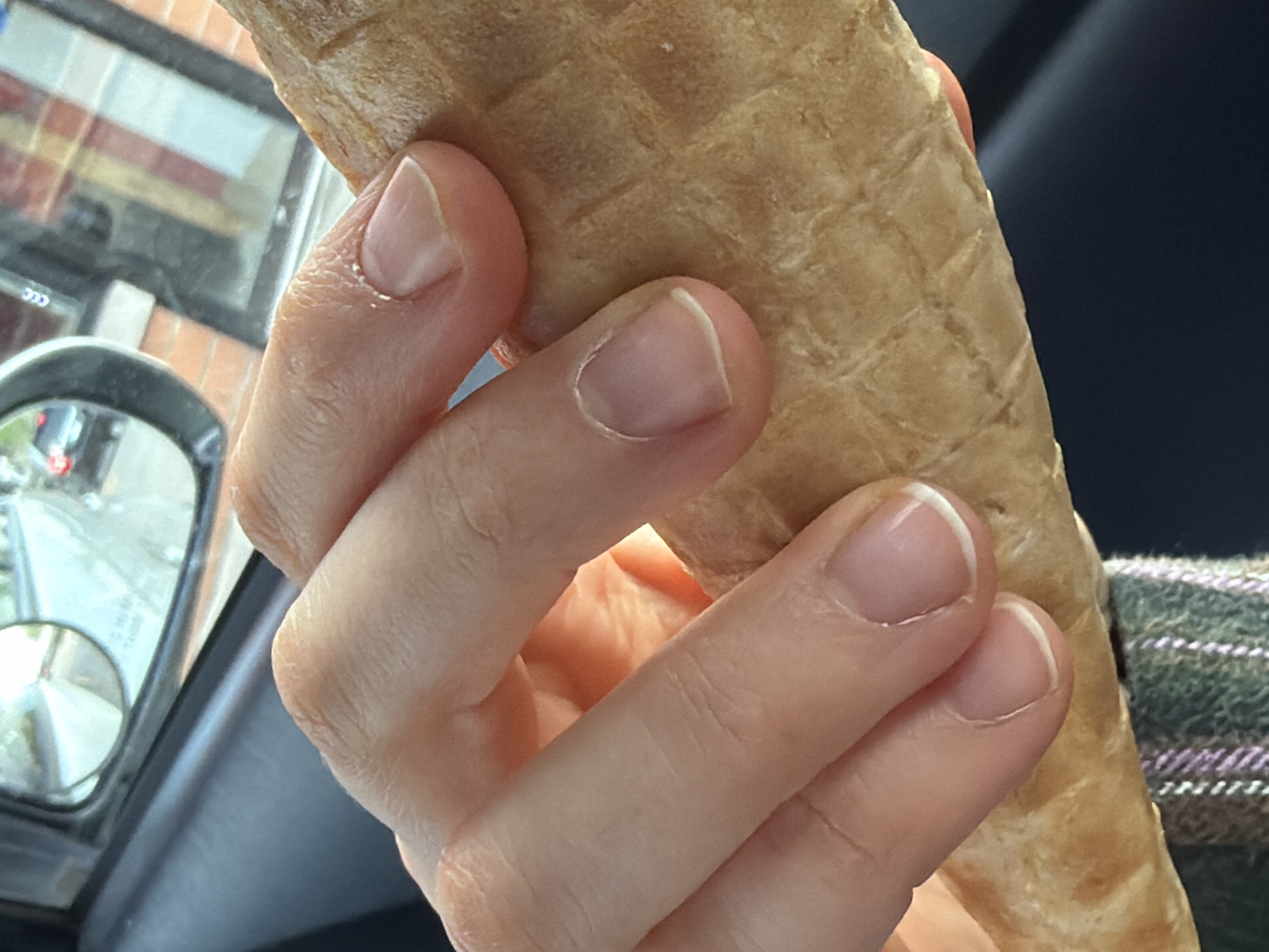 A close-up photograph shows a person's hand holding a waffle cone. The cone is light brown with a textured, grid-like pattern. The hand is fair-skinned with short, unpolished fingernails. The background includes a side mirror of a car, reflecting a street scene with buildings, possibly indicating an urban environment. The presence of a car interior and a street scene visible through the mirror suggest the picture was taken in a vehicle driving through or parked in an urban setting. Based on the environment, it is not possible to determine definitively that the picture was taken in Chicago. The reflection in the side mirror shows traffic, buildings and other elements that are associated with an urban setting and potentially a city in the United States.