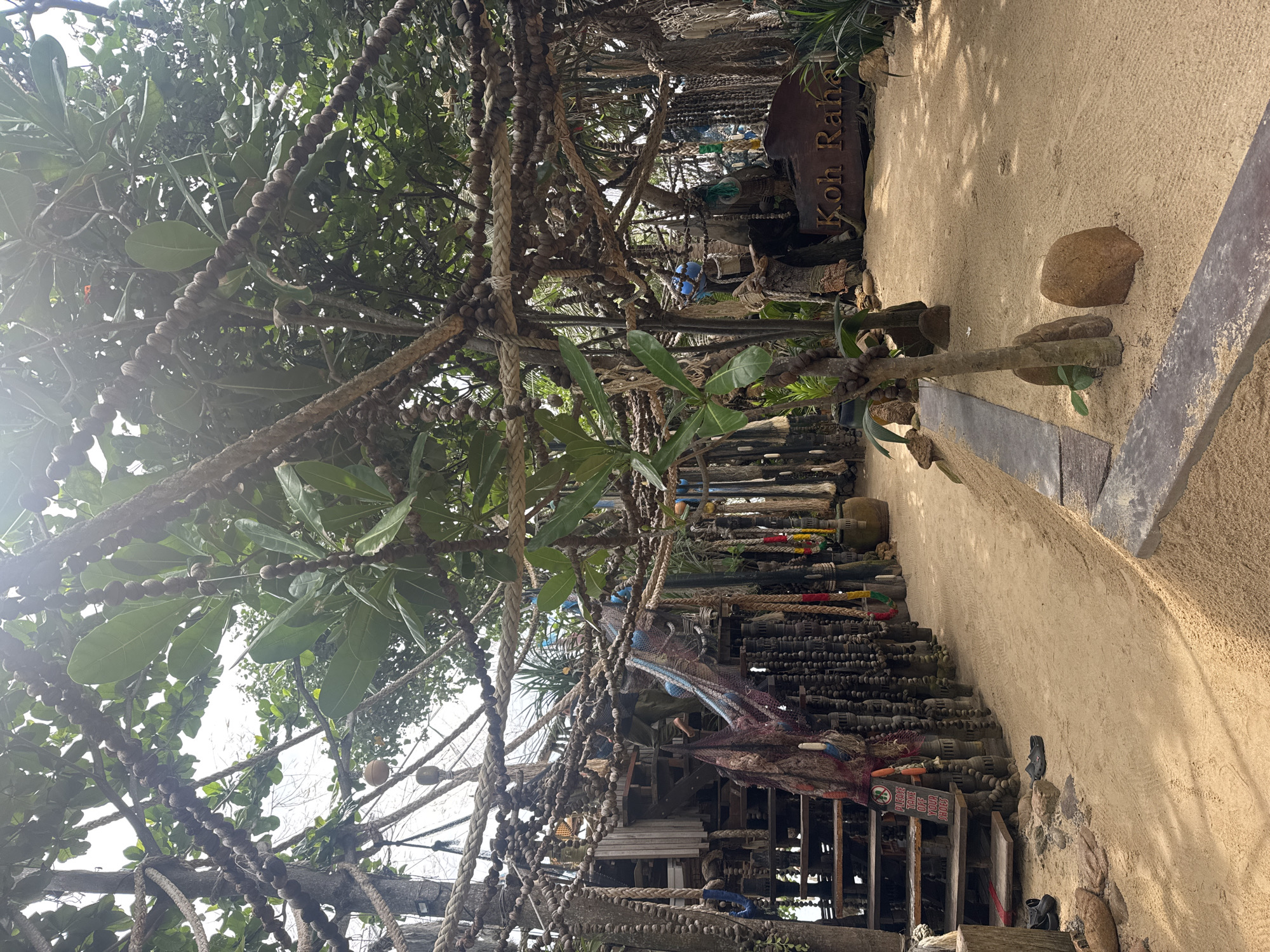 The image shows a path in ตำบลเกาะพะงัน, Thailand, with a sign reading "Koh Rahan" on the right. The path is sandy and bordered by a raised edge. The scene is heavily decorated with ropes adorned with spherical objects hanging from trees and structures above. Beneath the ropes, there is a series of steps leading to a platform decorated with various objects.