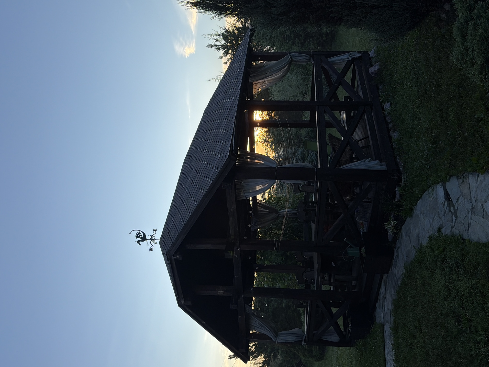 A wooden gazebo with a dark shingled roof stands on a grassy lawn. A dragon weather vane is mounted on the peak of the roof. The gazebo has open sides with curtains hanging from the eaves. Inside, tables and chairs are visible. A stone pathway leads to the gazebo. The scene appears to be set in a rural or suburban environment, possibly a backyard, at dusk. The sky is clear with a few wisps of clouds. The sun is setting behind trees in the background, casting a warm glow. The image is taken from a slightly low angle. The location is not specified as being in Leninsk-Kuznetskiy, Russia.
