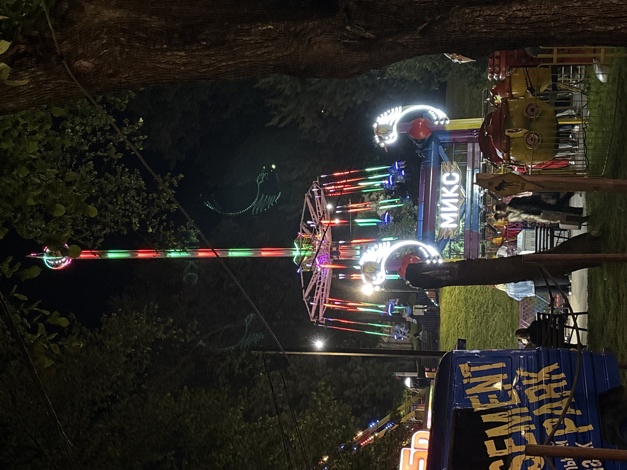This image depicts a nighttime scene at an amusement park, likely located in Vitsebsk, Belarus, given the Cyrillic signage. A tall amusement ride with a spinning carousel and brightly lit poles dominates the center of the frame, with red and green lights illuminating the structure. In the foreground, a blue van with yellow lettering that reads "CEMENT PARK" is visible. Several people are scattered throughout the park, some seated on benches and others walking. The park is surrounded by trees and a dark, night sky. Other amusement rides with colorful lights are visible in the background. The overall atmosphere is festive and lively, characteristic of a nighttime amusement park setting.