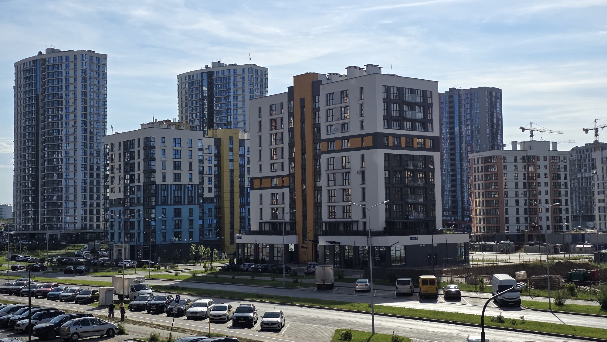 The image shows a modern residential complex in Minsk, Belarus. The scene is dominated by several tall, multi-story apartment buildings with contemporary architectural designs, featuring balconies and large windows. The buildings display a variety of colors and materials, including white, grey, blue, and orange accents. In the foreground, a street is visible with several parked cars and a few individuals walking. Several more cars are parked in lots adjacent to the buildings. Construction cranes are visible in the background, suggesting ongoing development in the area. The sky is partly cloudy, indicating daytime. The overall impression is of a developing urban landscape.