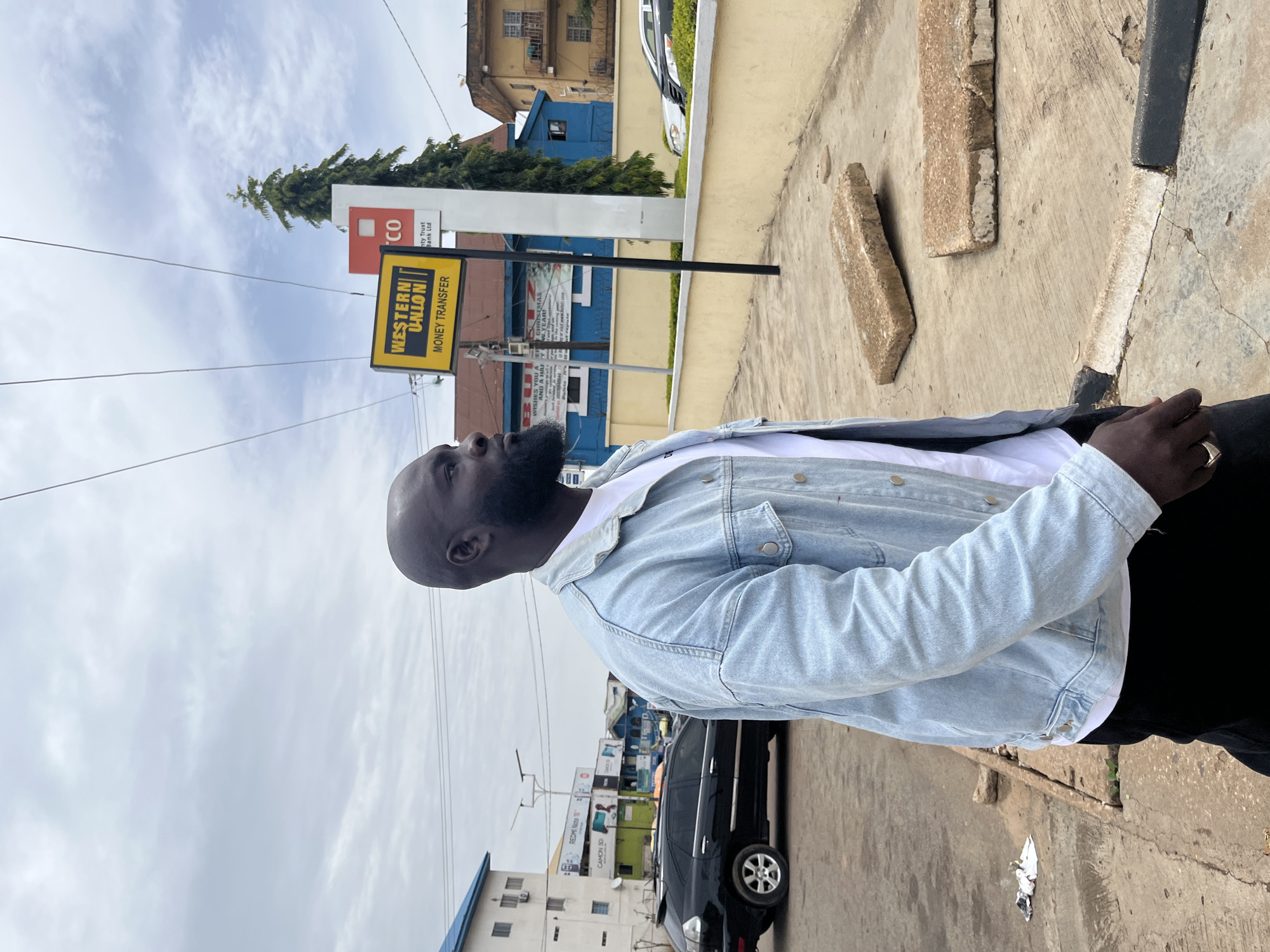 A bald, dark-skinned man wearing a light blue denim jacket and a white t-shirt stands facing right, looking towards the distance. He has a beard and is wearing a ring on his right hand. He is positioned on a concrete sidewalk in front of a low, beige wall. Behind the wall, a blue building with a pitched roof is visible, along with a tall evergreen tree. To the left of the blue building, a yellow sign advertising "WESTERN UNION MONEY TRANSFER" is mounted on a pole. Further left, a black SUV is parked. Buildings and streetlights are visible in the background. The sky is overcast with clouds. The scene appears to be in an urban environment.