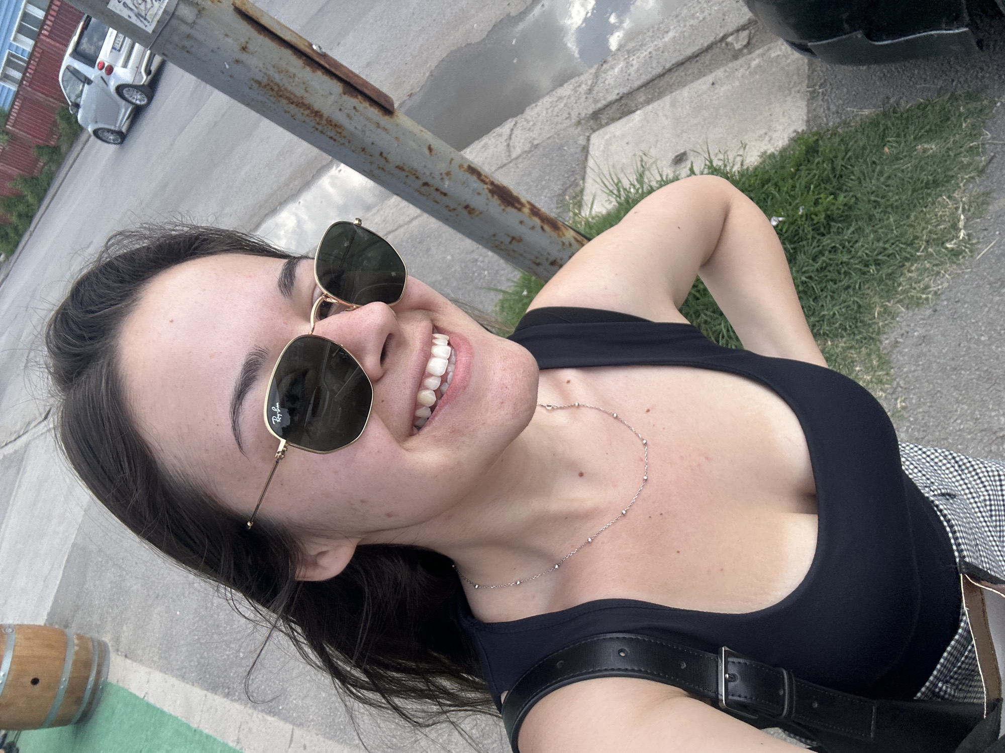 The image shows a selfie of a woman wearing a black tank top, sunglasses, and a necklace. She is standing outdoors with a street and a grassy area in the background. A wooden barrel is visible to the left, and a lamppost is to the right. A silver car is parked on the street in the distance. The woman is smiling and her hair is blowing in the wind. The location is identified as being in the city of Sofia, Bulgaria.