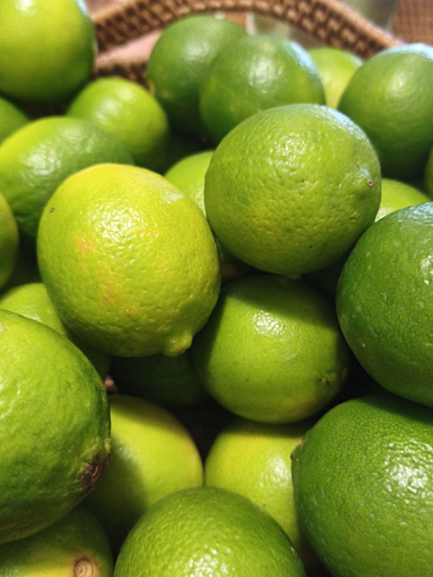 A close-up, overhead shot captures a pile of ripe, green limes. The limes are round and vary in size, with some exhibiting subtle variations in color and texture, including a few with small brown blemishes. The focus is sharp on the foreground limes, with a shallow depth of field blurring the background, where hints of a woven basket can be seen. The lighting is bright, highlighting the glossy surfaces of the fruit. The scene suggests a market or harvest setting. The image does not contain any information that would allow for the determination of a specific location in Bac Nha Trang Ward, Vietnam, or the presence of any people, actions, or interactions.