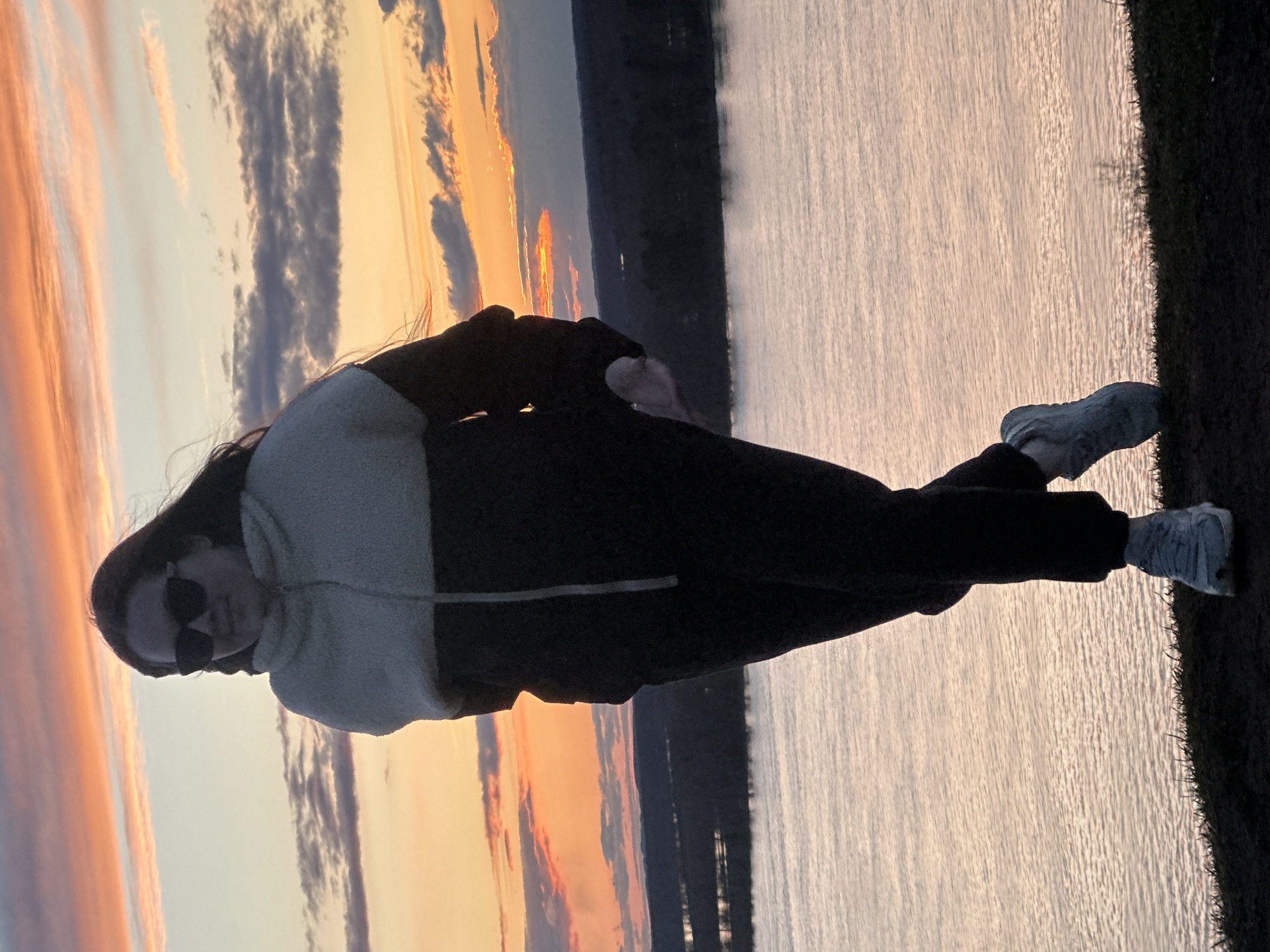 The image, when viewed with the horizon oriented horizontally, depicts a full-body shot of a person standing outdoors against a dramatic sunrise or sunset backdrop. The subject, appearing to be female, is positioned on a grassy or earthy bank overlooking a wide body of water, likely a river or lake. Across the water, a dark, distant landmass or shoreline is visible under a vast sky.
The person is wearing dark sunglasses, a two-tone jacket—light-colored on the upper section and dark on the lower—dark trousers, and light-colored athletic shoes. Her long, dark hair flows down her back. She is standing slightly angled, with her right arm bent across her body.
The sky is a notable feature, displaying a spectacular blend of warm hues including oranges, pinks, and yellows, intermingled with soft blues and greys, punctuated by scattered clouds. The low sun, peeking above the distant horizon, creates a bright glow that reflects warmly on the rippling surface of the water, contributing to the strong backlighting of the scene. The foreground and the subject are partially silhouetted by this light.
The scene captures a tranquil moment in nature, with the individual seemingly observing or enjoying the scenic view during the golden hour. No visible text is present in the image. The location context for this serene landscape is Ольховка, Russia.