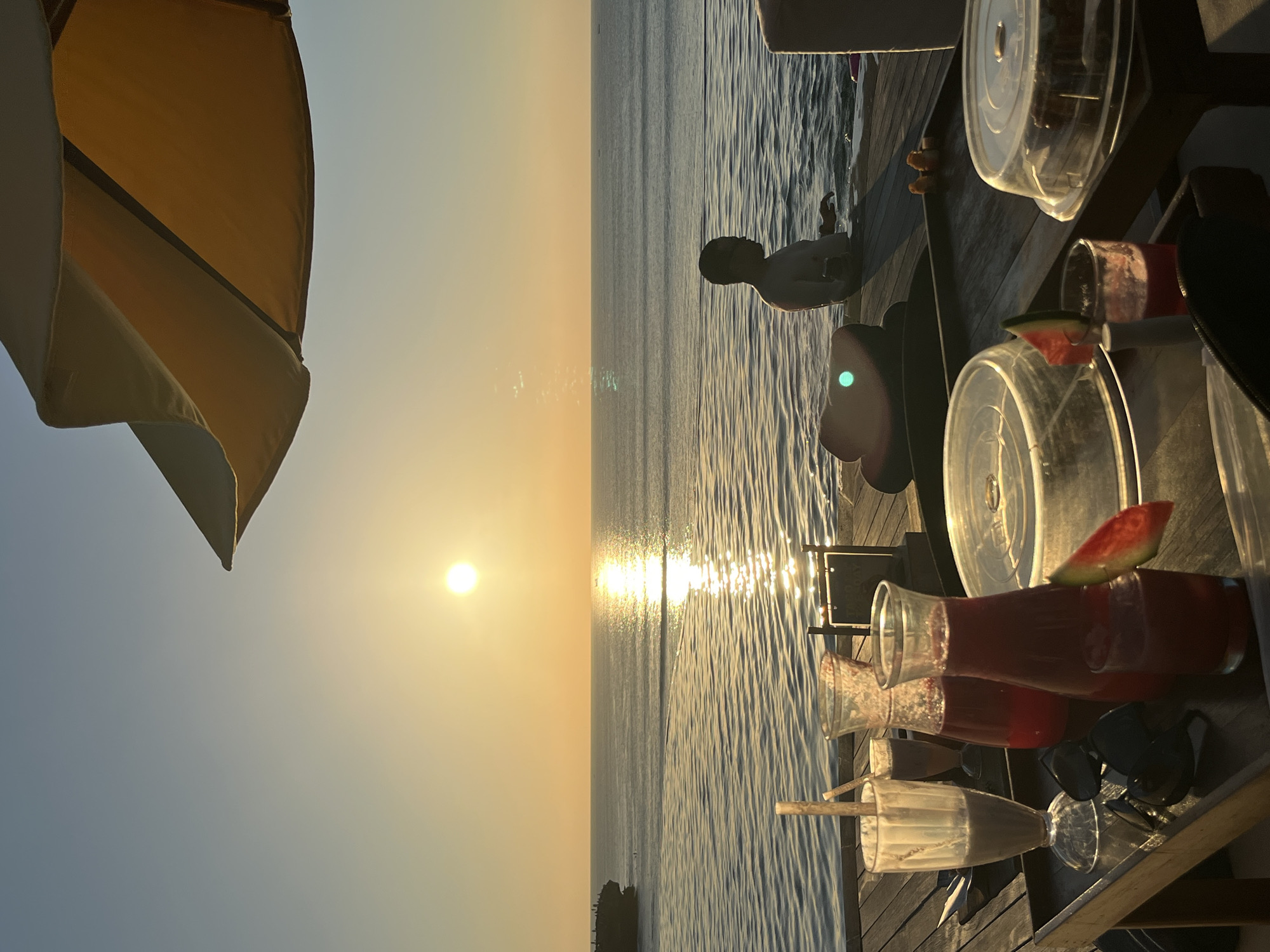 At sunset on a wooden deck overlooking the ocean, a man is partially submerged in an infinity pool. The sky is a gradient of pale blue and warm orange, with the sun a bright orb near the horizon. The ocean reflects the sun's golden light. In the foreground, a tray holds several drinks, including a glass of chocolate-colored liquid with a straw, two carafes filled with a red beverage, and a small glass with a piece of watermelon garnish. Sunglasses and a slice of watermelon are also on the tray. To the right, a cake covered with a clear dome is visible. The scene appears to be set at a resort in Kuta Selatan, Indonesia, given the architectural style of the deck and the surrounding tropical ambiance.