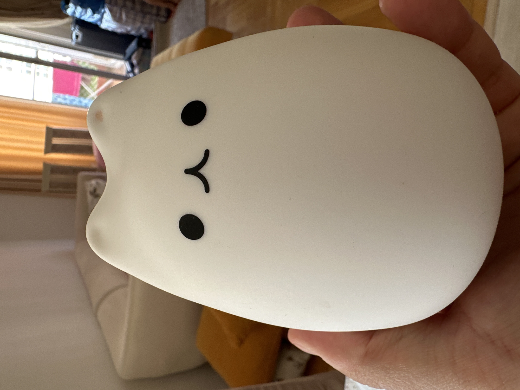 A white silicone cat-shaped night light is being held in a person's hand. The night light has black dot eyes and a curved line mouth. The background is blurred, but appears to be an indoor setting with furniture. The lighting suggests it is daytime. There is a small brown discoloration on the upper left side of the cat's head.