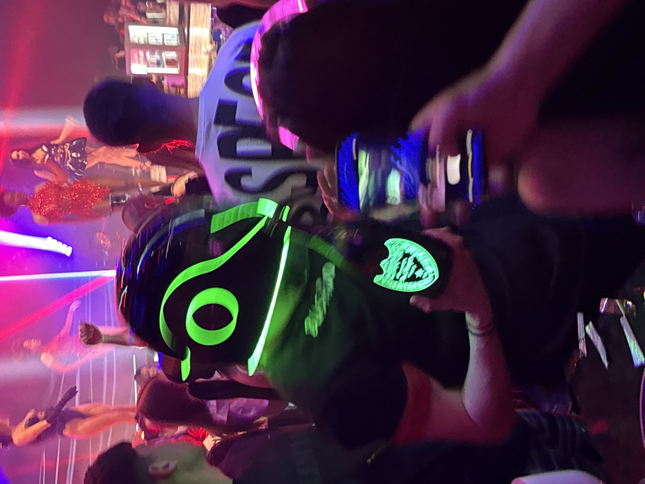 The scene depicts a crowded nightclub interior in Plovdiv, Bulgaria, characterized by dynamic, multicolored stage lighting in pink, purple, and blue hues, and atmospheric smoke. In the foreground, a person wears a black helmet or headpiece with prominent neon green glowing outlines and a circular motif, along with a black top featuring glowing green text that appears to read "BUSTER." This individual holds a dark bottle with a distinctly glowing neon green Dom Pérignon shield logo. To the immediate right, another person is partially visible, holding a smartphone with a bright blue illuminated screen, seemingly capturing an image or video of the event. In the midground and background, numerous other attendees are present, with various figures observing the stage. On an elevated stage or platform in the background, several female performers are visible: one in a dark, form-fitting costume with long gloves; another in a light-colored, possibly sheer dress; a third in a short, shimmering red dress; and a fourth in a patterned short dress. One performer is observed with a raised fist. The overall environment is dark and vibrant, typical of a high-energy entertainment venue.