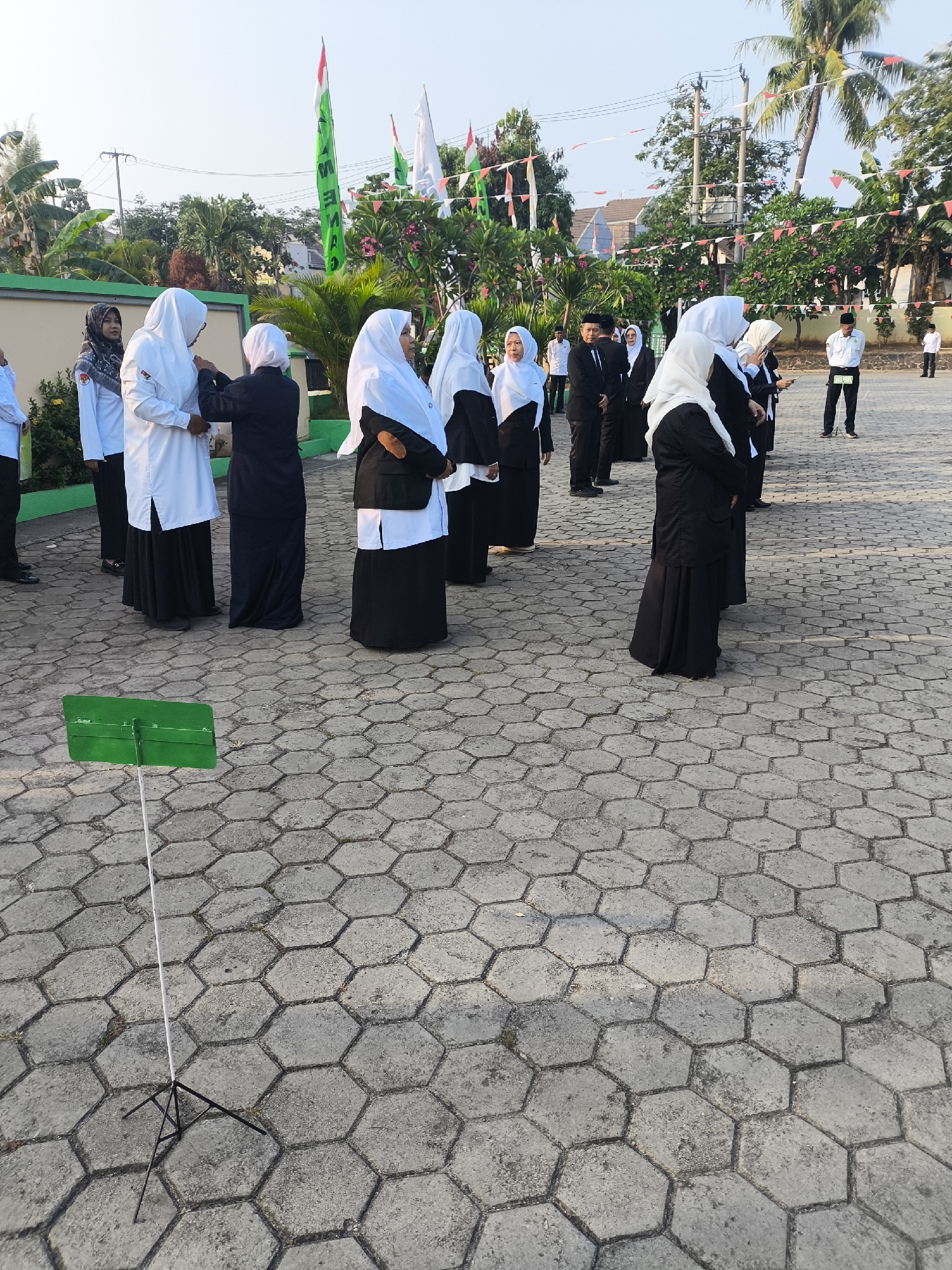 The image captures an outdoor event with several individuals, primarily women wearing white and black hijabs and attire, standing in a formation on a hexagonal paver stone surface. Some individuals are wearing white shirts and black skirts or pants, while others are in black attire. In the background, there are palm trees, buildings, and several flags, including Indonesian flags (red and white) and green flags. There is also a green music stand with a white pole in the foreground. The scene appears to be in a public space, possibly a school or community gathering, given the uniformed attire and flags. The location is specified as Cilegon, Indonesia.