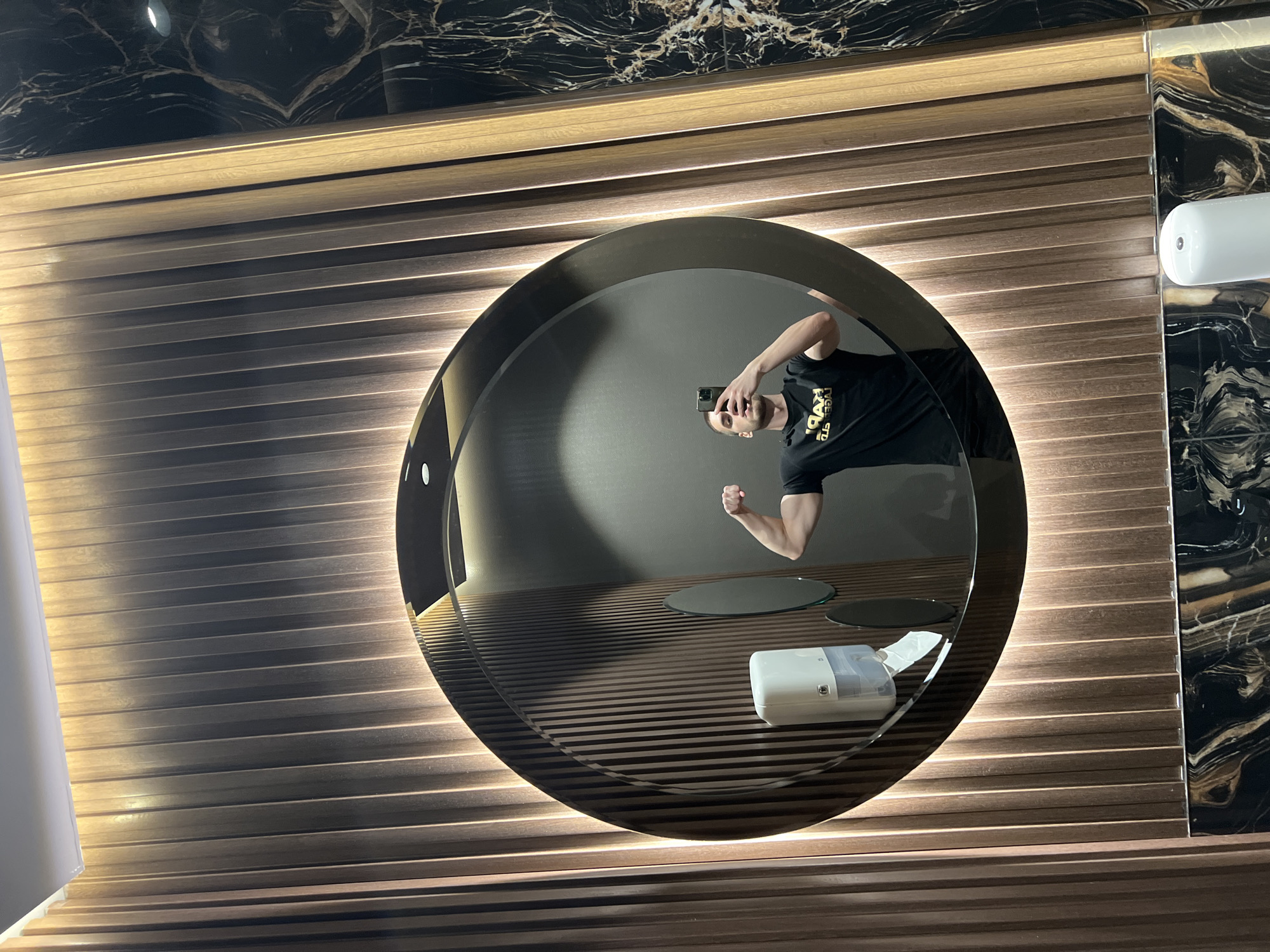 The image captures a modern interior, likely a restroom or changing room, presented from a rotated, sideways perspective. A large, circular mirror is the central focus, featuring a backlit frame that emits a warm, soft glow. Reflected in the mirror is a man with short hair and a beard, taking a selfie with a smartphone. He wears a dark t-shirt with text that, when un-reflected, appears to read "ROYAL STANDARD." His free arm is bent, showcasing a flexed bicep in what seems to be a playful pose.
Inside the mirror's reflection, a white rectangular dispenser, possibly for soap or tissues, is visible on a surface, accompanied by two small, dark circular reflective discs. The wall surrounding the mirror is clad in horizontal slatted panels, illuminated from behind to create glowing linear patterns. Above and to the right of these slats, the wall transitions to dark marble or stone tiles with prominent gold and white veining. A white cylindrical light fixture is partially visible on the far right. The scene is illuminated by artificial light, emphasizing the contemporary design and sleek finishes. The location is Burgas, Bulgaria.