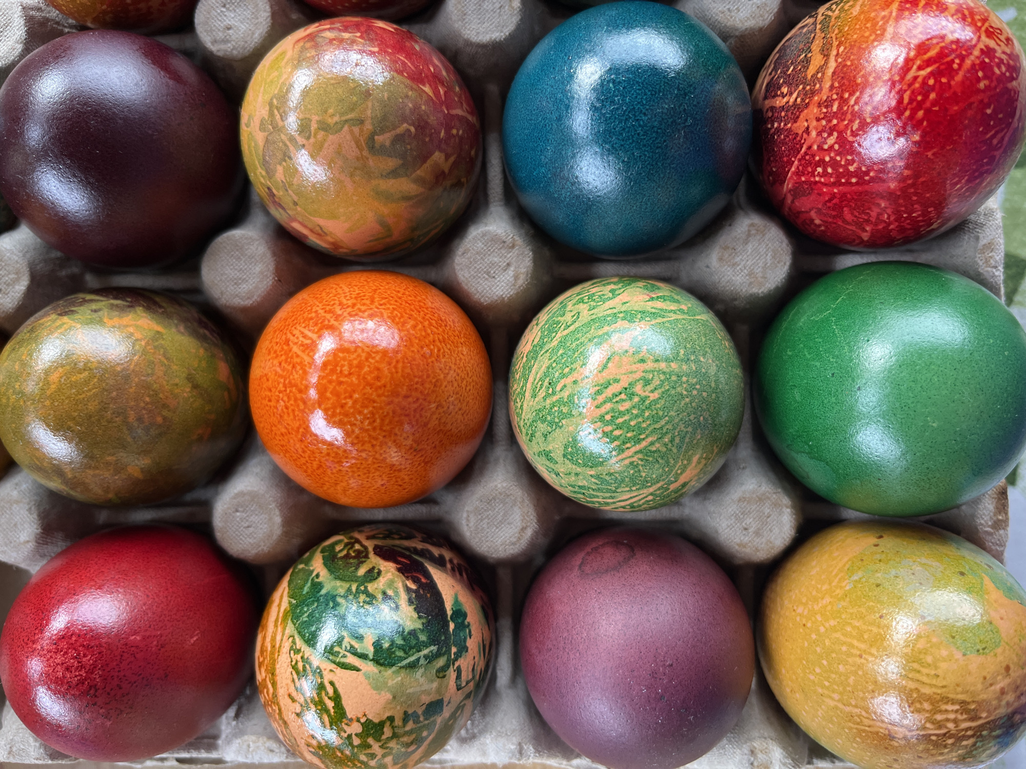 A close-up overhead shot shows twelve brightly colored Easter eggs nestled in a cardboard egg carton. The eggs are arranged in a 3x4 grid. The colors vary, including shades of red, orange, yellow, green, blue, and purple. Some eggs have mottled or marbled patterns. The carton is neutral in color and appears to be made of molded pulp. The eggs are shiny, suggesting they may have been oiled after dyeing. The shot is presumably taken in Pchelnik, Bulgaria, but there's no way to confirm this from the image itself. There are no people or interactions visible in the frame.