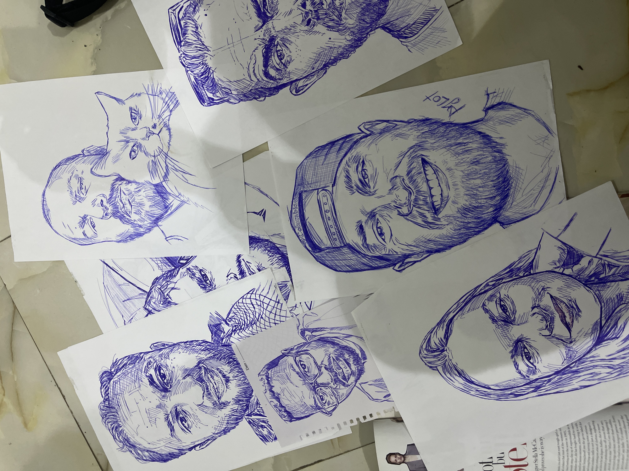 The image shows several blue ink pen portraits drawn on white paper, scattered across a light-colored, textured floor that appears to be indoors. The portraits feature men, one woman, and a cat. One portrait depicts a man holding a cat. The style of the drawings is sketch-like, with visible cross-hatching and linework. The overall impression is that of an artist's workspace or a collection of completed sketches. The floor's texture suggests it could be tiles or concrete, common in residential or commercial spaces. No specific location within Abuja, Nigeria, can be identified from the visual information provided.
