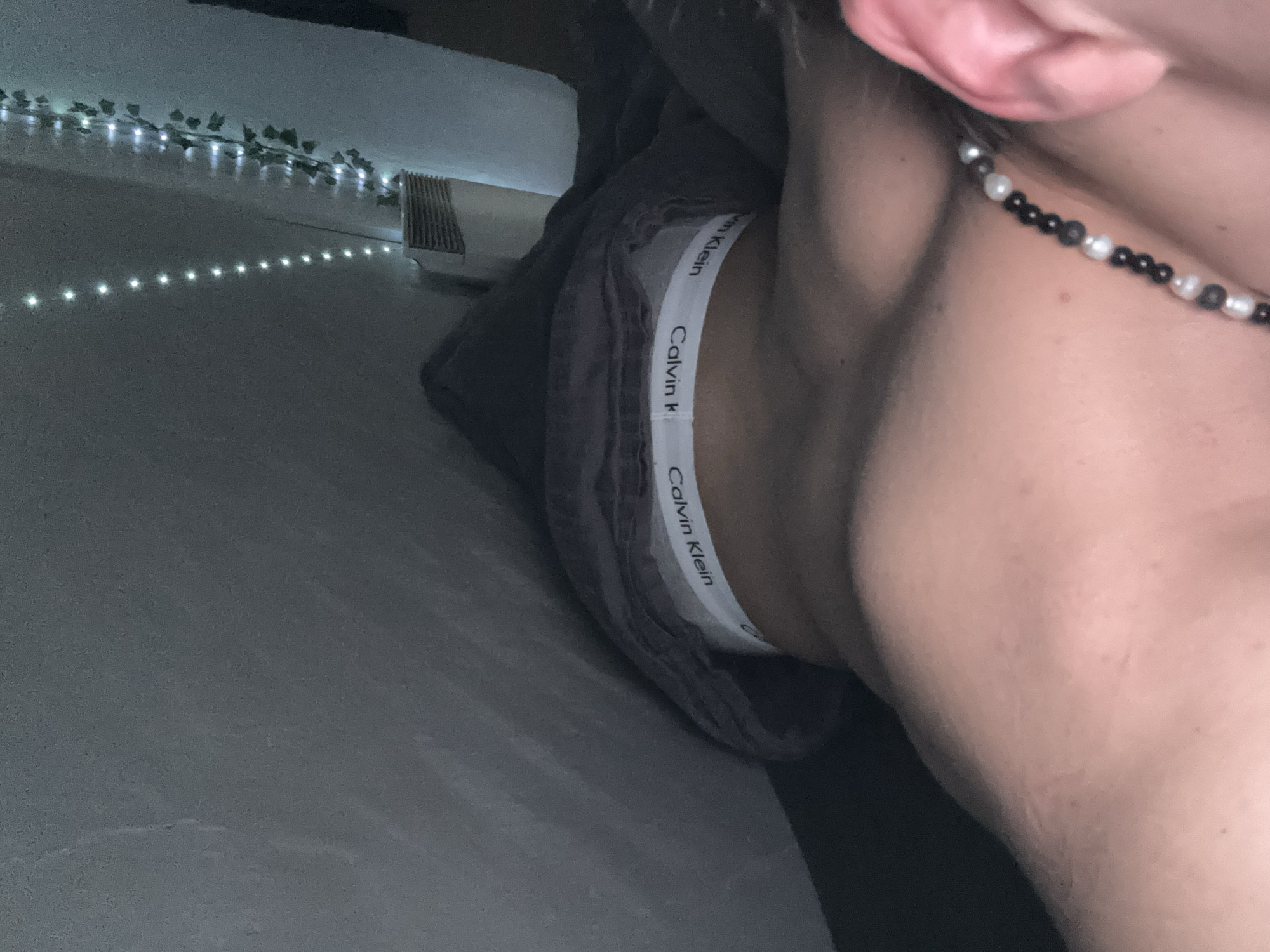 The image shows a close-up view of a person's neck and upper torso. The person is wearing a Calvin Klein underwear band. A necklace with black and white beads is visible. String lights are in the background, with some green leaves interspersed along the string. A radiator is partially visible near the lights. The environment appears to be indoors, possibly a bedroom. The image does not contain information to identify a location in the city of Troyan, Bulgaria.