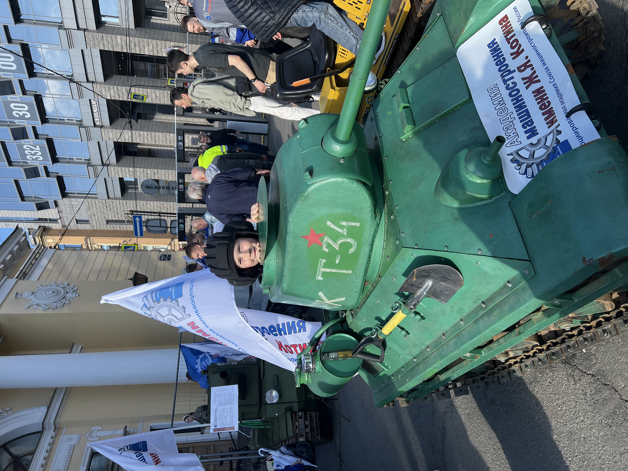 The image shows a green, vintage T-34 tank parked on a city street. A person wearing a padded helmet is visible in the tank's hatch. Red star emblem and "K T-34" are written on the tank. A shovel is attached to the front of the tank. A sign on the tank is in Russian and refers to the "Academy of Mechanical Engineering named after J.Ya. Kotin." There are multiple flags with mechanical engineering symbols behind the tank. Several people are standing around the tank, looking at it. A building with glass windows and the word "CEHATOP" is visible in the background. Numbers 330 and 332 are on the corner of the building. Based on architectural style and the Cyrillic text, it's likely located in Saint Petersburg, Russia.
