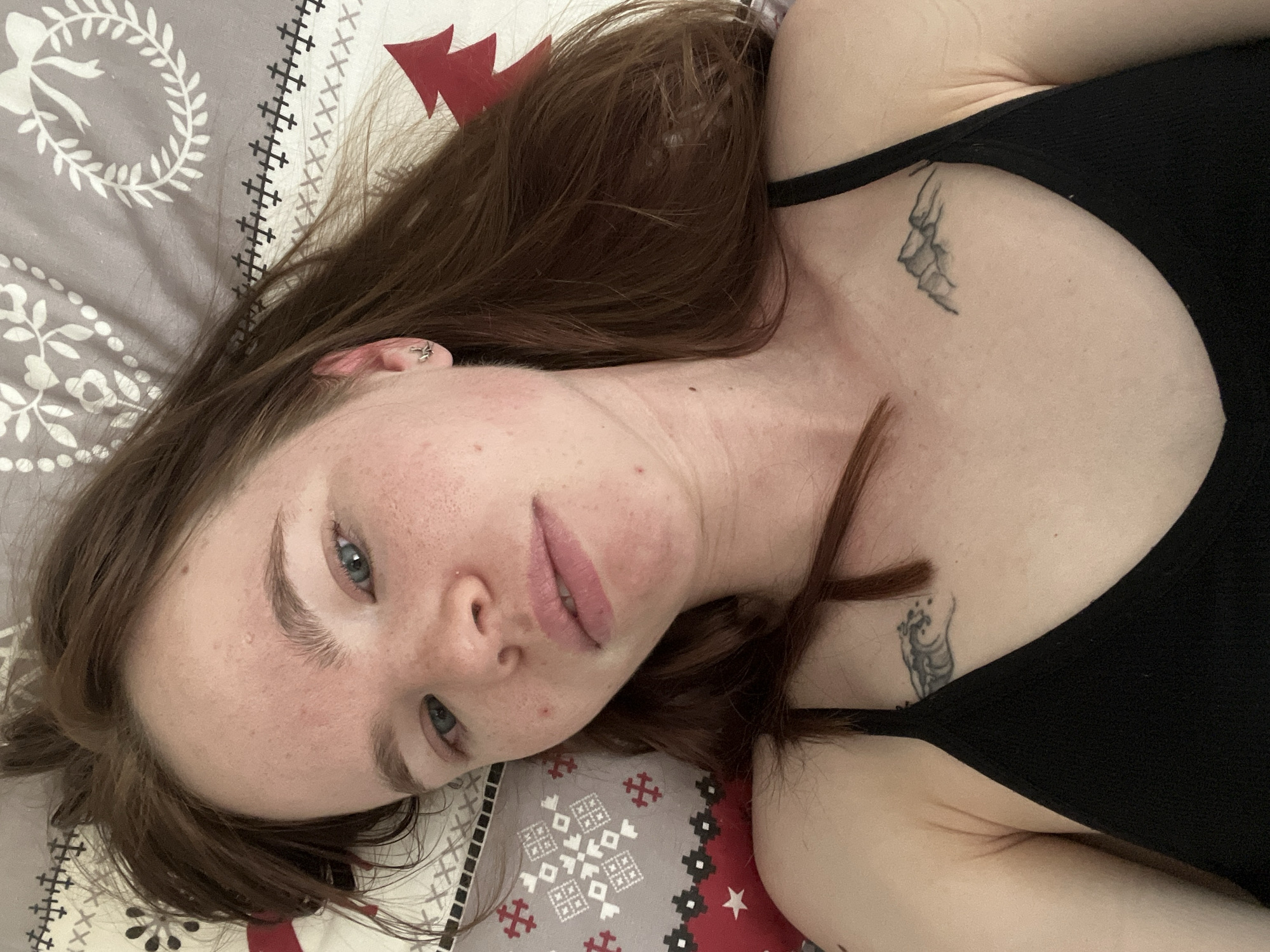 A selfie shows a woman with fair skin, freckles, and light eyes, lying on a patterned bedsheet. She has reddish-brown hair. She is wearing a black tank top. Tattoos are visible on her upper chest. The bedsheet features geometric patterns in shades of gray, red, and white. The location is unknown, and there is no indication that the image was taken in Perm, Russia.