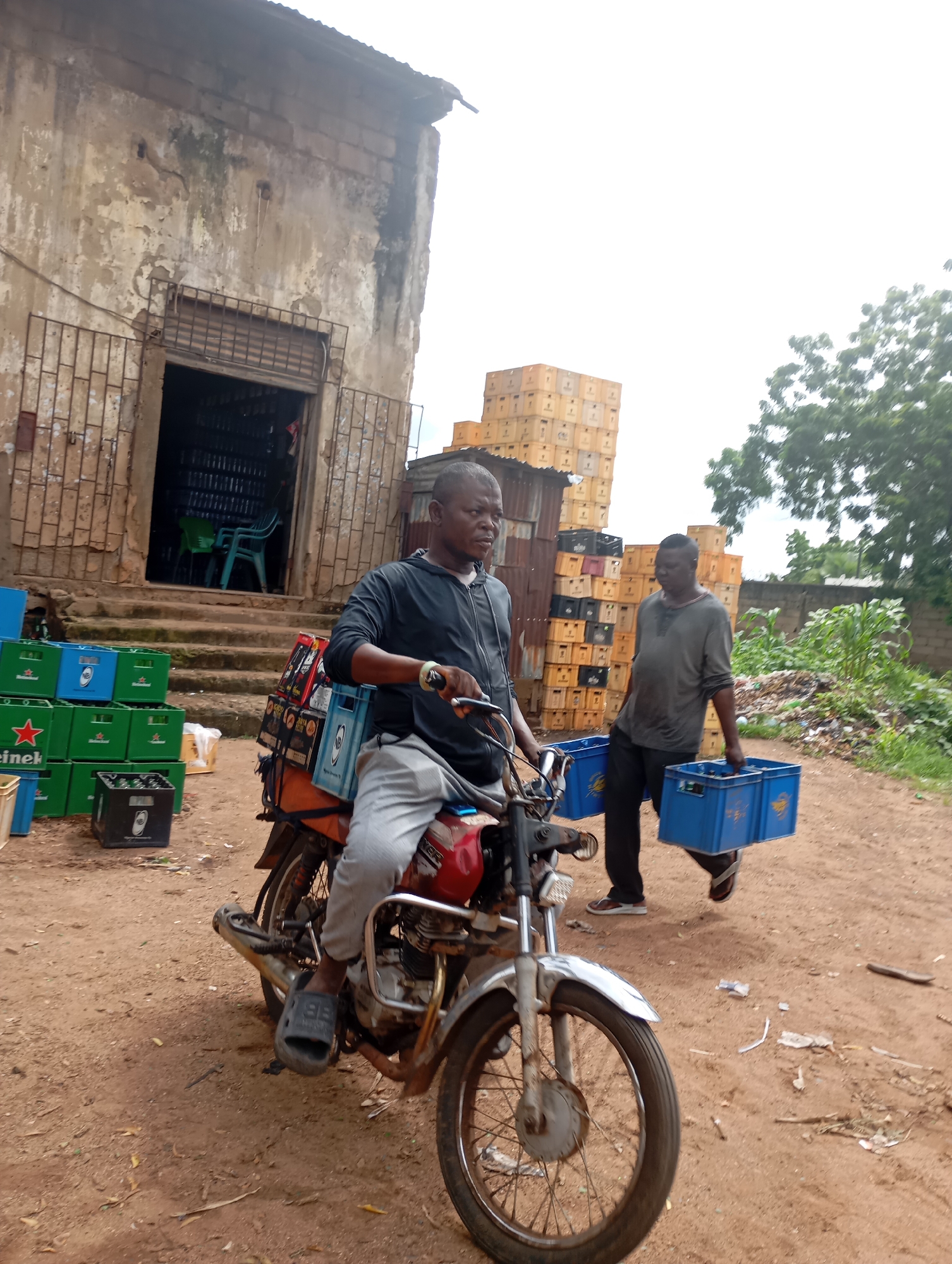 In Shendam, Nigeria, an outdoor scene depicts two men involved in the distribution of beverages. The foreground features a man riding a motorcycle laden with cases of drinks, likely beer, strapped to the back. He is wearing a black long-sleeved shirt and grey pants, with black footwear. Beside him, to the right, a second man walks, carrying two blue plastic crates, each filled with drinks. He is dressed in a grey long-sleeved shirt and black pants, with sandals.
The environment suggests a commercial area with a dilapidated building in the background. The building has a weathered facade, a metal grate over its entrance, and steps leading up to it. To the left of the building, stacks of green and blue plastic crates, many bearing branding that appears to be "Heineken," are visible. Behind the men and to the right, a large accumulation of stacked yellow crates dominates the background, interspersed with some darker crates. Trees and dense vegetation are present in the far background, and the ground is covered in reddish-brown earth, scattered with debris. The overall atmosphere suggests a busy, perhaps informal, distribution hub.
