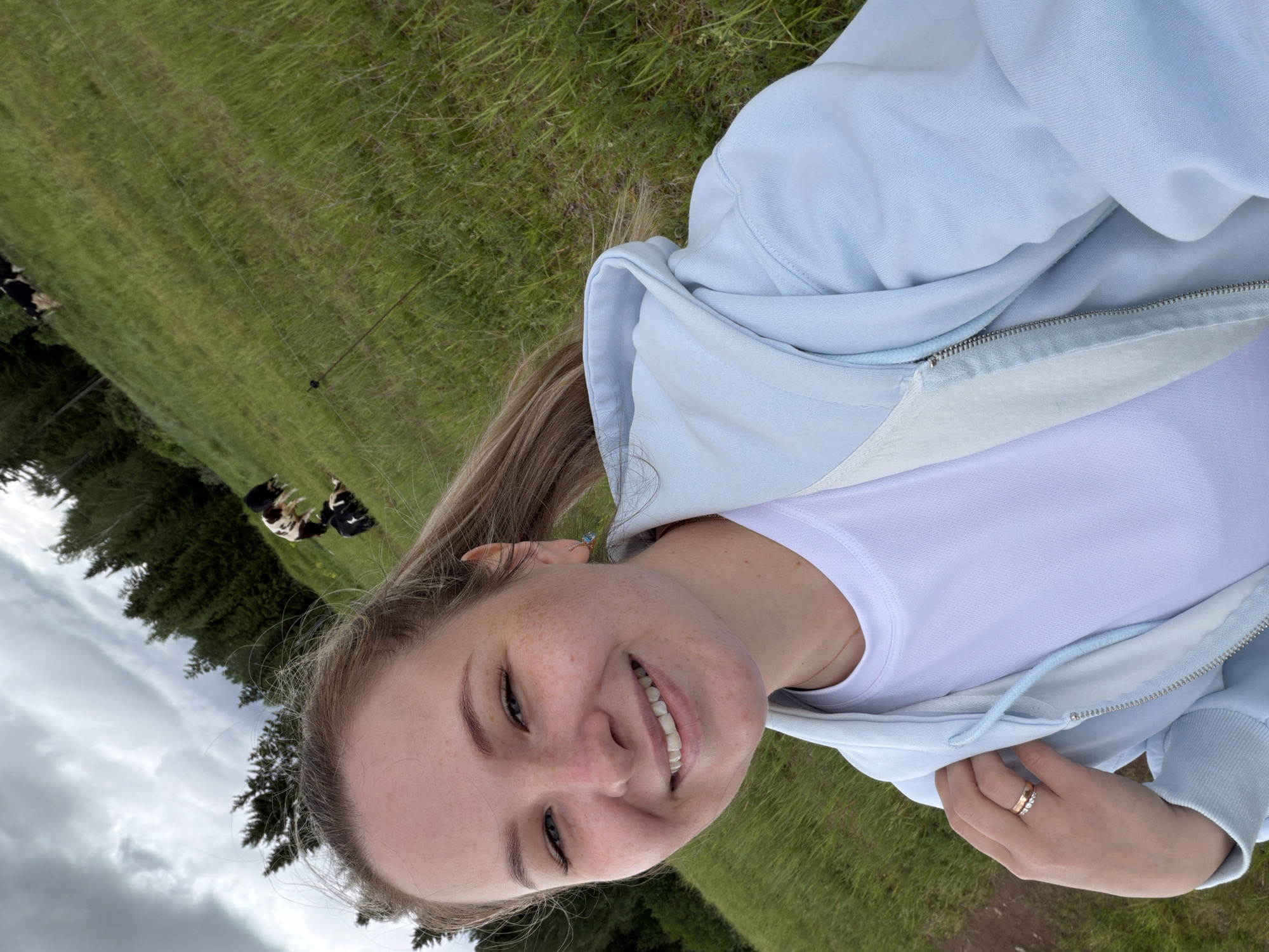 A young woman smiles into the camera while wearing a light blue zip-up hoodie and a white t-shirt. Her blonde hair is pulled back, and she has fair skin with freckles. She wears rings on her right hand. The background shows a grassy field with a treeline on the left. A few cows are visible in the field. Based on the location inferred from the query, this scene might be situated in the rural areas surrounding Ольховка, Russia, known for its farmland and natural landscapes.