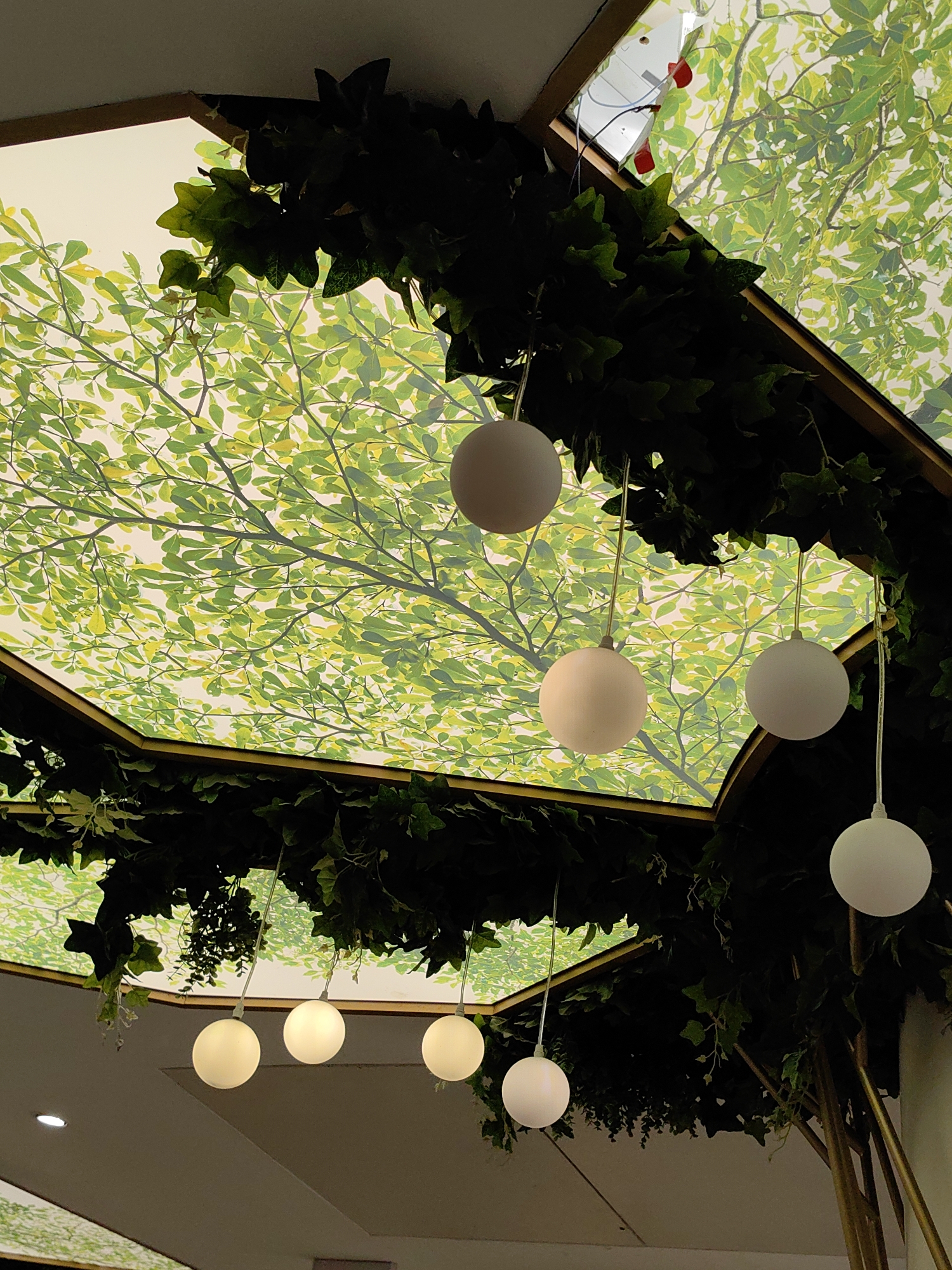 The image shows a decorative ceiling with a canopy of artificial green leaves and hanging spherical lights. The ceiling itself appears to be a printed mural of a tree canopy with bright green leaves and branches. The lighting fixtures are simple white spheres suspended by thin wires. The overall impression is one of an artificial, indoor garden ambiance. There are no people visible in the image. The scene is likely from an indoor public space, possibly a restaurant, cafe, or a shopping mall, designed to evoke a natural environment. The location is specified as Mumbai, India.