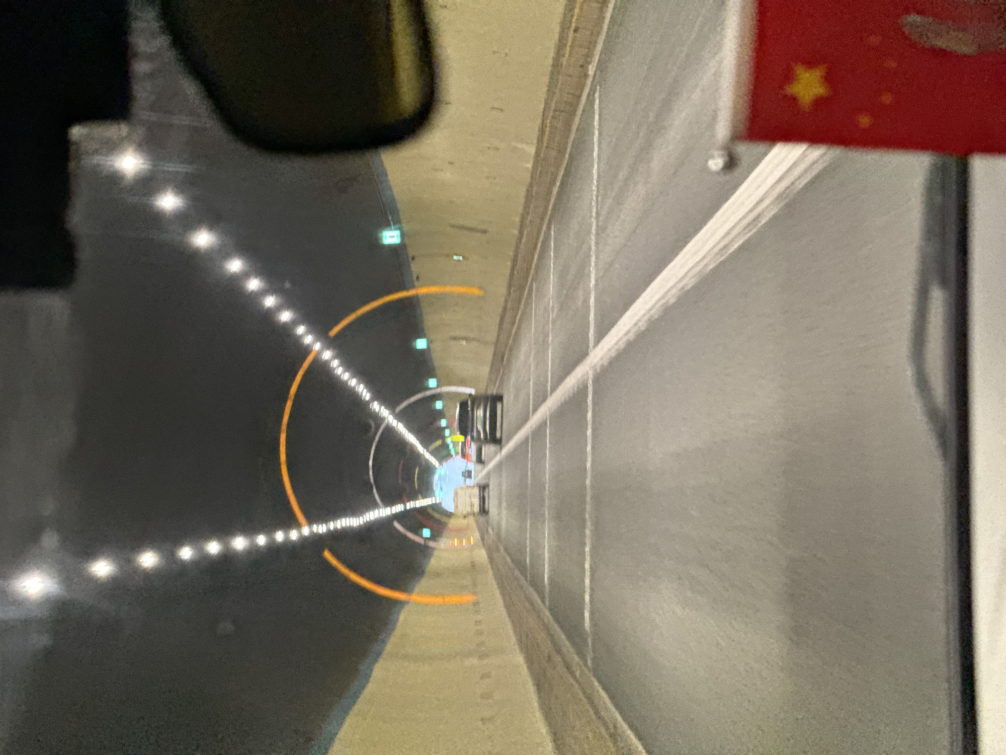 The image captures a forward-facing view from inside a moving vehicle within a multi-lane road tunnel in Xiehe, China. The tunnel features light-colored concrete walls and a dark ceiling illuminated by sequential white strip lights forming an arc overhead and extending down the tunnel. Orange-lit archways are visible at intervals, complementing the primary lighting. Green rectangular lights are positioned along the upper walls. The asphalt road surface displays clear white lane markings. Ahead, a white utility van, a dark passenger car, and a black SUV or sedan are visible, all traveling in the same direction. A portion of a rearview mirror is visible in the upper right, and a small red flag with a yellow star, consistent with the flag of the People's Republic of China, is partially visible in the bottom right corner, suggesting its attachment to the vehicle's interior.