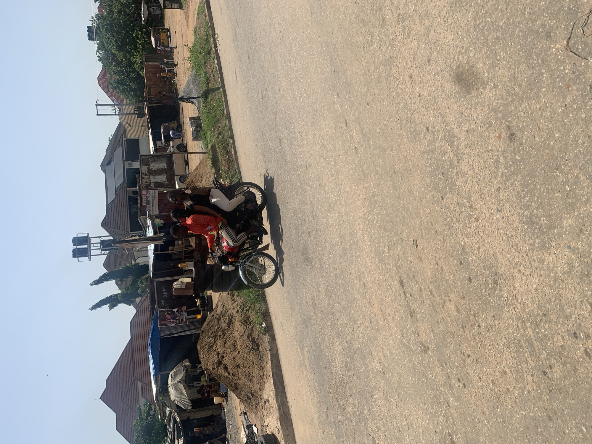 The image depicts a street scene in Abuja, Nigeria. A motorcycle carries two people. The person in the front wears a red shirt, while the rear passenger wears white trousers. They are on a paved road. To the left of the road, there is a line of buildings and structures, including shops and residences. There's also a pile of dirt or sand near the roadside. Overhead utility lines are visible. The sky is light blue, indicating daylight.