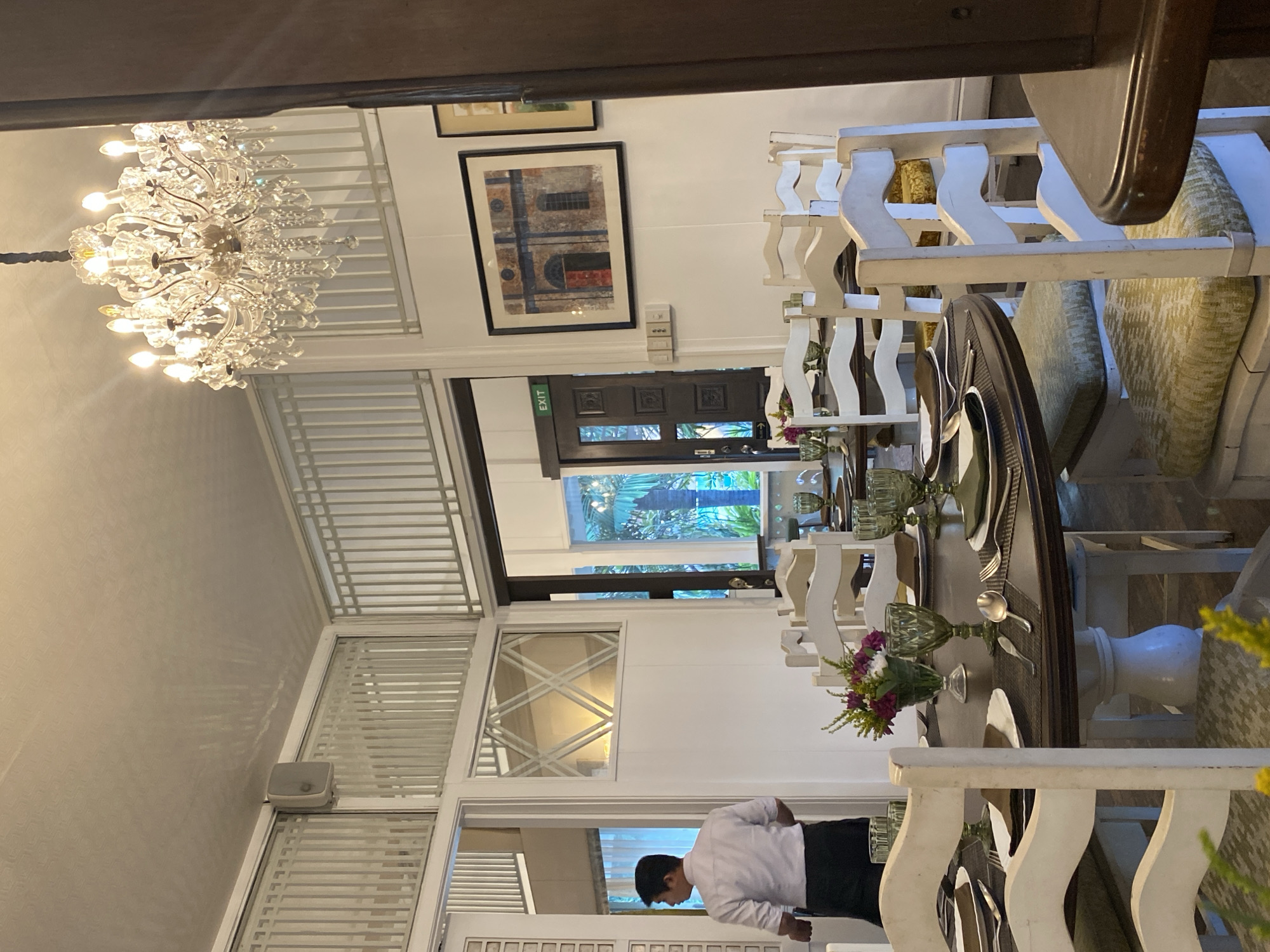 This is a media file showing an indoor dining scene, possibly a restaurant. The primary subjects are the dining setup and a person. The scene is brightly lit, suggesting it might be daytime, though artificial lighting from a chandelier is also prominent.
The setting appears to be a well-decorated indoor space with white-paneled walls and elegant white furniture, including chairs with ornate backs and a large, dark-wood dining table. The table is set with plates, glassware, and silverware, indicating a meal service is either in progress or being prepared. A decorative chandelier hangs from the ceiling. Artwork is displayed on the walls, and a large window or doorway offers a glimpse of greenery outside, possibly a garden or courtyard.
A person, dressed in a white shirt and dark trousers or apron, is visible walking in the background, facing away from the camera. Their posture suggests they are attending to their duties in the dining area.
Notable details include the detailed craftsmanship of the chandelier and the furniture. The presence of place settings suggests an event related to dining, such as a meal or a special occasion. The location cues, such as the style of architecture and the hint of tropical greenery outside, along with the provided location context of Davao City, Philippines, suggest a warm climate.
There is a visible "EXIT" sign in green text on a dark door frame. Other minor text or signage may be present on unseen elements.