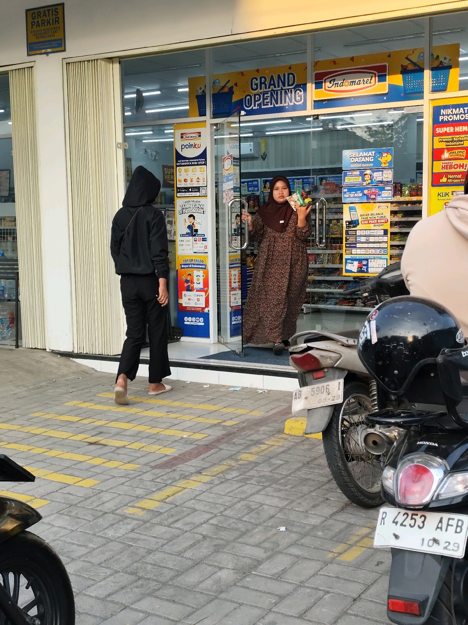 In Sanden, Indonesia, at an Indomaret convenience store, a woman wearing a brown hijab and a patterned dress stands at the entrance, holding several small packaged items. To her left, a person in a black hooded jacket and black pants walks away from the store. In the foreground, two motorcycles are parked on a paved surface with yellow painted lines. The store's facade features signage indicating a "Grand Opening" and "Indomaret" branding. The environment appears to be an outdoor commercial area during daylight.
