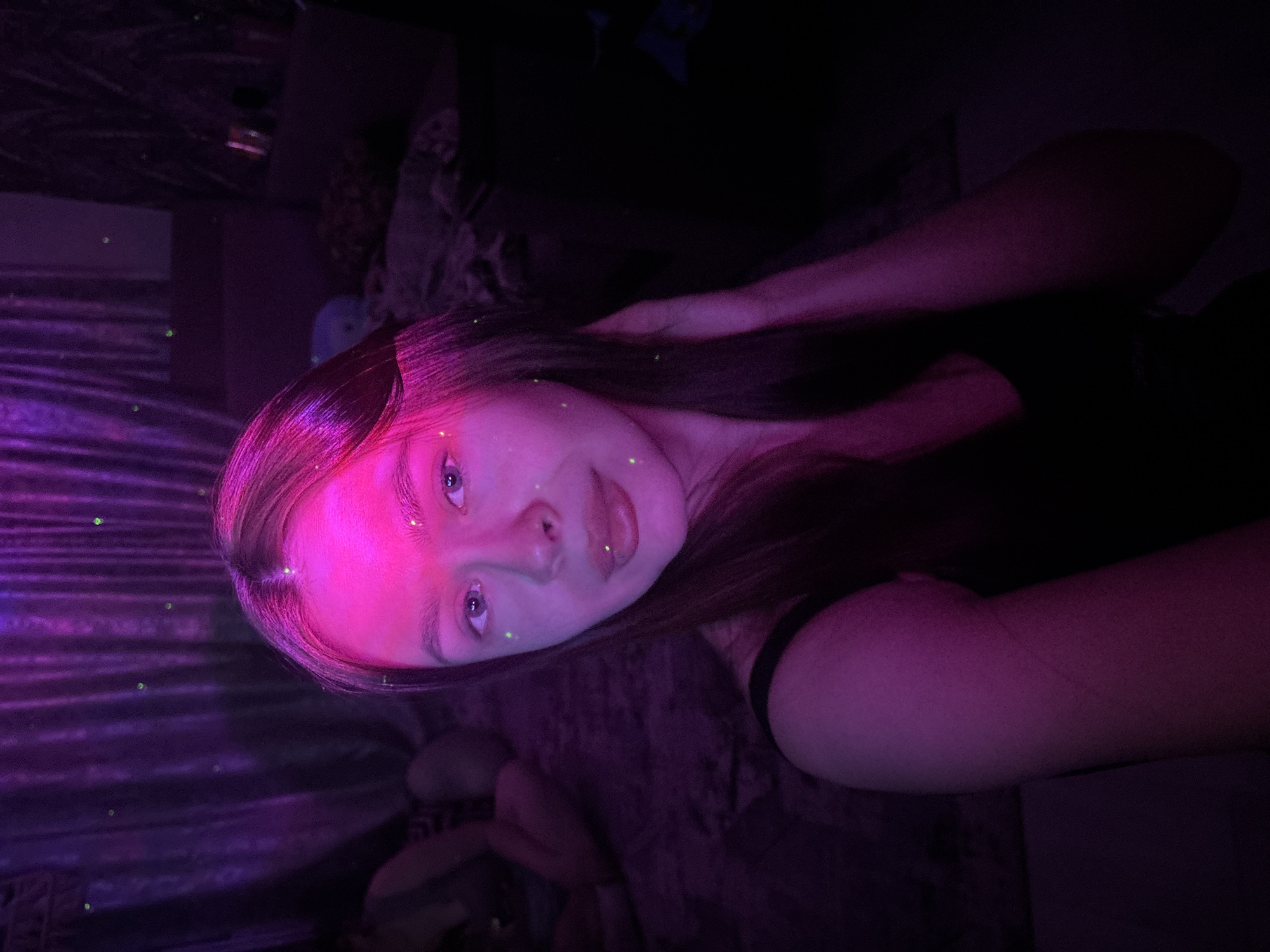 This image depicts a vertical-format selfie of a light-skinned female, approximately in her late teens or early twenties, positioned centrally in a dimly lit indoor setting. She has long, dark brown hair and is looking directly into the camera with a neutral expression and slightly parted lips. A prominent pink light source illuminates her forehead, nose, and the upper part of her right cheek, casting a magenta glow. Numerous small, bright green circular light points are scattered across her face (forehead, right cheek, chin), hair, and the background. She is wearing a black or dark-colored sleeveless top. Her left arm and hand are visible, with the hand raised to touch her hair near her right temple.
The background shows an interior space, heavily shadowed, but illuminated by the same ambient pink and scattered green lights. To the upper left, a purple, pleated curtain or fabric drape is visible, featuring several green light points. Below this, partially obscured and blurred, are the lower legs and feet of another individual, positioned horizontally on a textured, dark surface, likely a bed or floor. The remainder of the background to the right of the primary subject is obscured by darkness, with only faint, indistinct shapes suggesting furniture or objects. The specific location within the city of Chaikovsky, Russia, cannot be determined from the visual information presented in the image.