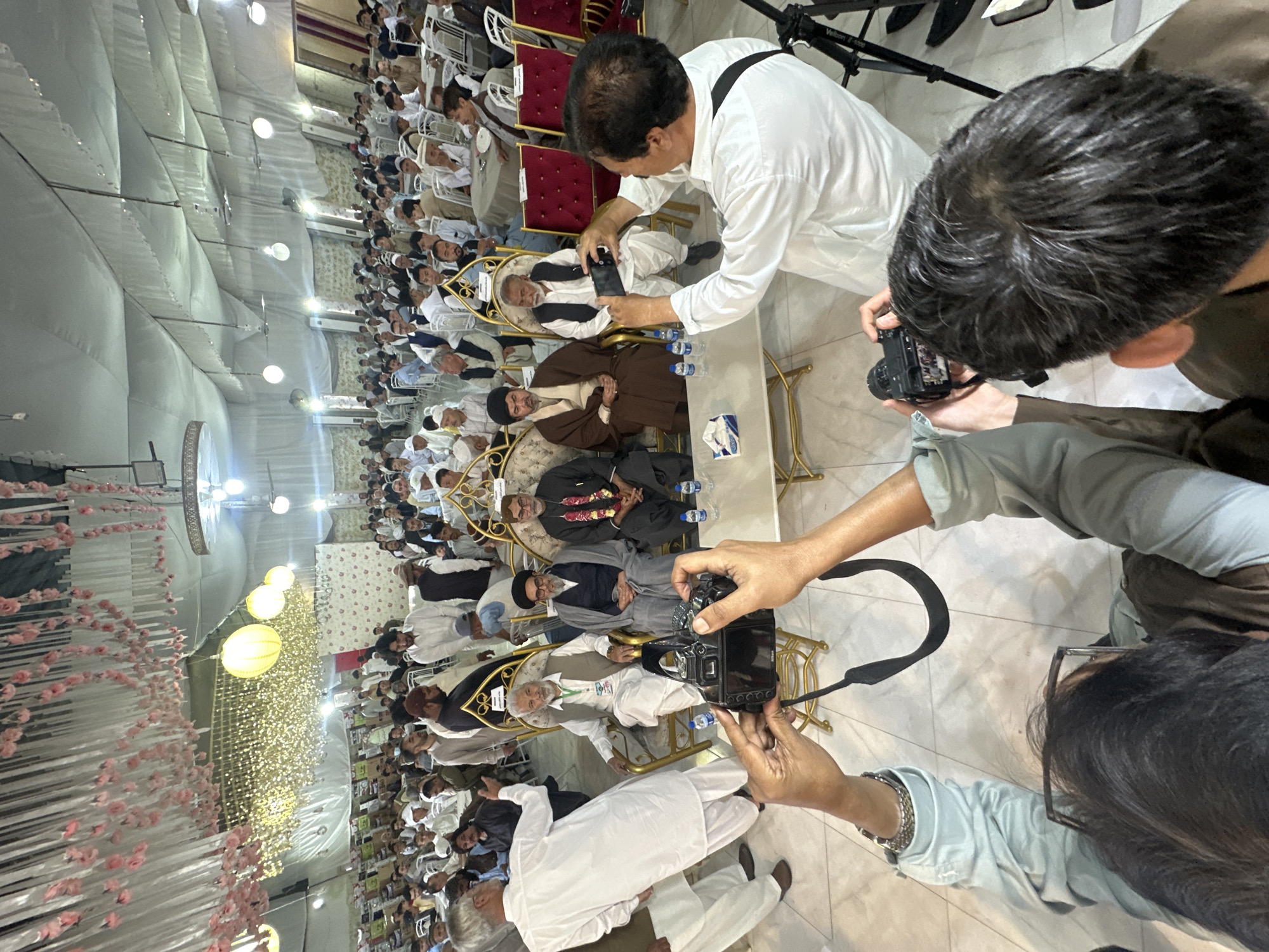 The image depicts an event in Quetta City Tehsil, Pakistan, where multiple men are seated on golden chairs. Several people are taking photographs of the seated men using both cameras and mobile phones. The setting appears to be indoors under a large, white tent with decorative lighting. Long rows of men in white clothing are seated behind the main group. Water bottles are visible on a table in front of the seated men.