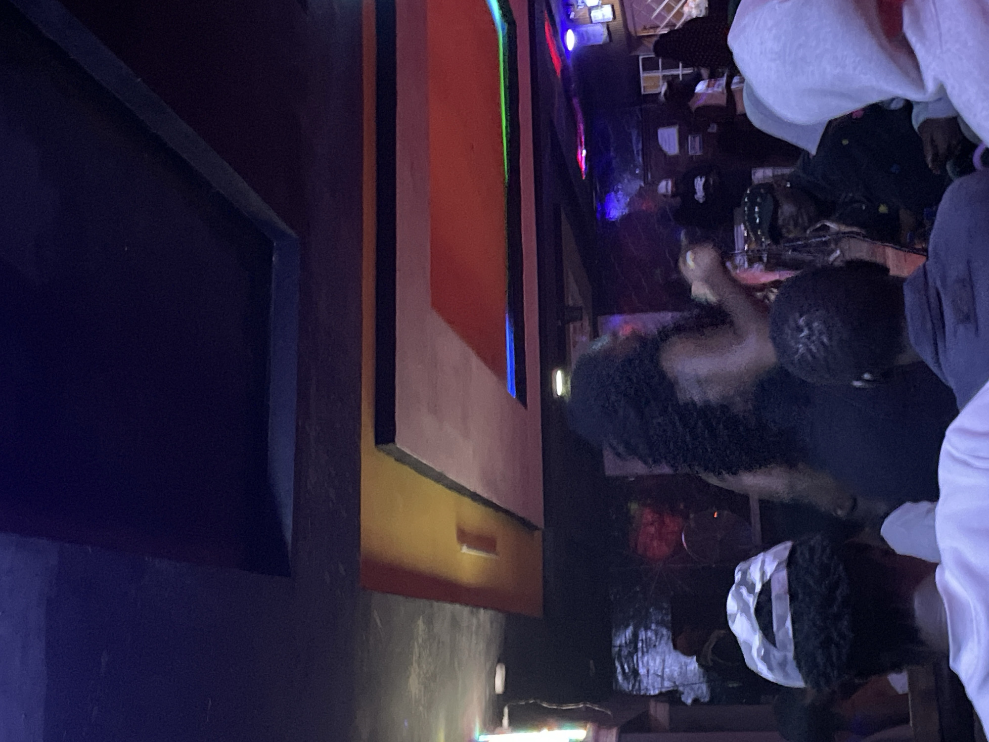 The image depicts an indoor scene at night, likely a club or entertainment venue, with a focus on a woman dancing in the center. The environment features a dark ceiling with recessed lighting and a layered ceiling design with colorful lighting accents, including red, orange, green, and blue, suggesting a vibrant atmosphere. In the foreground, several people are visible, with their backs to the camera or partially turned. One person wears a white baseball cap, another a grey hoodie, and others are in darker clothing. The woman in the center has long, dark, curly hair and is wearing a dark outfit. Her pose suggests she is actively dancing or performing. In the background, other individuals are visible, some appearing to be seated, and the overall lighting is dim, with the colorful lights from the ceiling creating a dynamic and somewhat blurred effect. The image quality is low, with a grainy appearance typical of low-light photography. The scene is consistent with an informal gathering or performance space within Durumi, Nigeria.