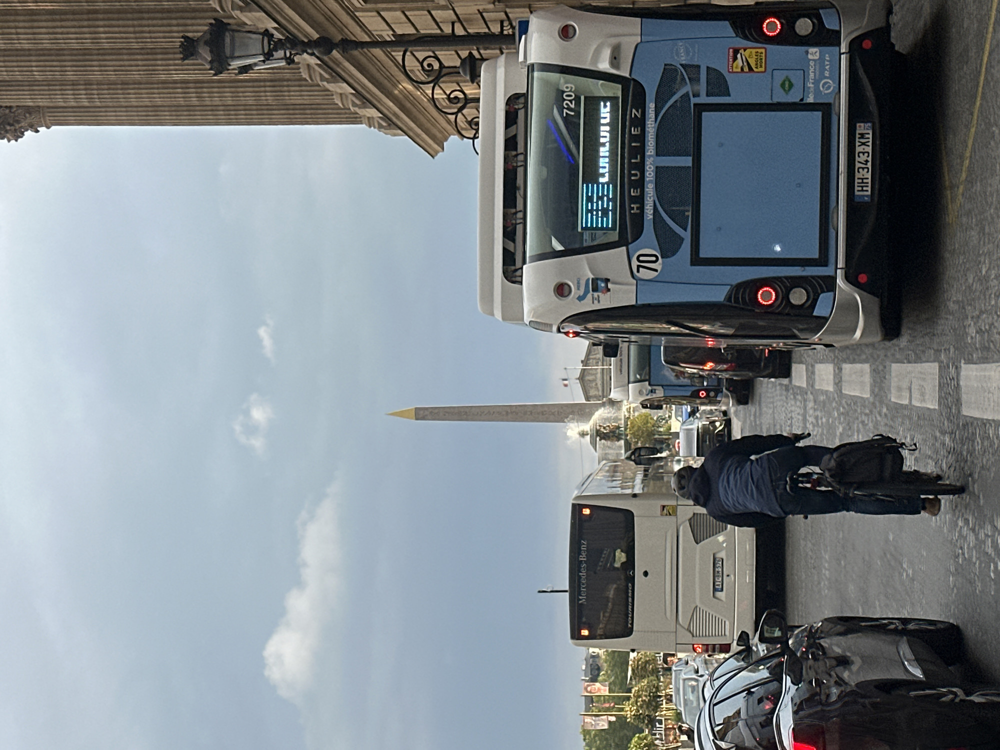 The image shows a street scene in Paris, France, featuring traffic and the Luxor Obelisk in the background. Two buses, one a Mercedes-Benz Tourismo and the other a HEULIEZ "véhicule 100% biométhane" are visible. The license plate of the HEULIEZ bus is HH-343-XM. The buses are surrounded by other vehicles. A cyclist rides on the road, dodging the traffic. The sky is partly cloudy. A building with columns and a lamppost is on the upper left of the image.