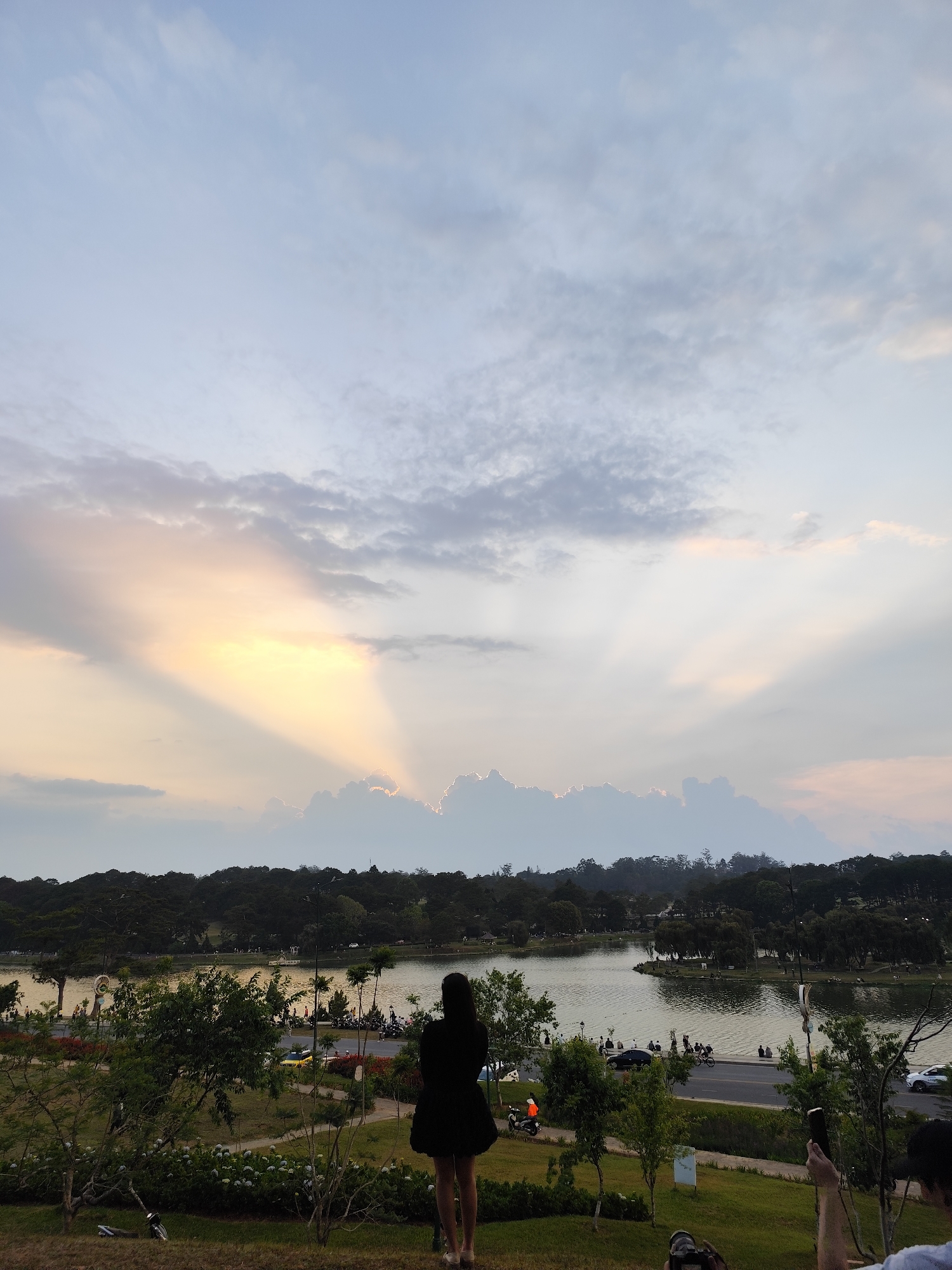 The image presents an outdoor scene in Da Lat, Vietnam, near Xuan Huong Lake, capturing the interplay of nature and human presence under an impressive sky.
In the foreground, a woman in a black dress stands on a grassy slope. To the right, a hand is seen holding a cell phone; and to the left, there is a bicycle.
Beyond the immediate foreground, a road stretches along the lake's edge, where people are walking, sitting, and riding scooters and cars.
Across the lake, dense trees create a forest canopy. In the background, a dramatic sky dominates the scene, featuring a combination of clouds and sunbeams emanating from behind the trees.