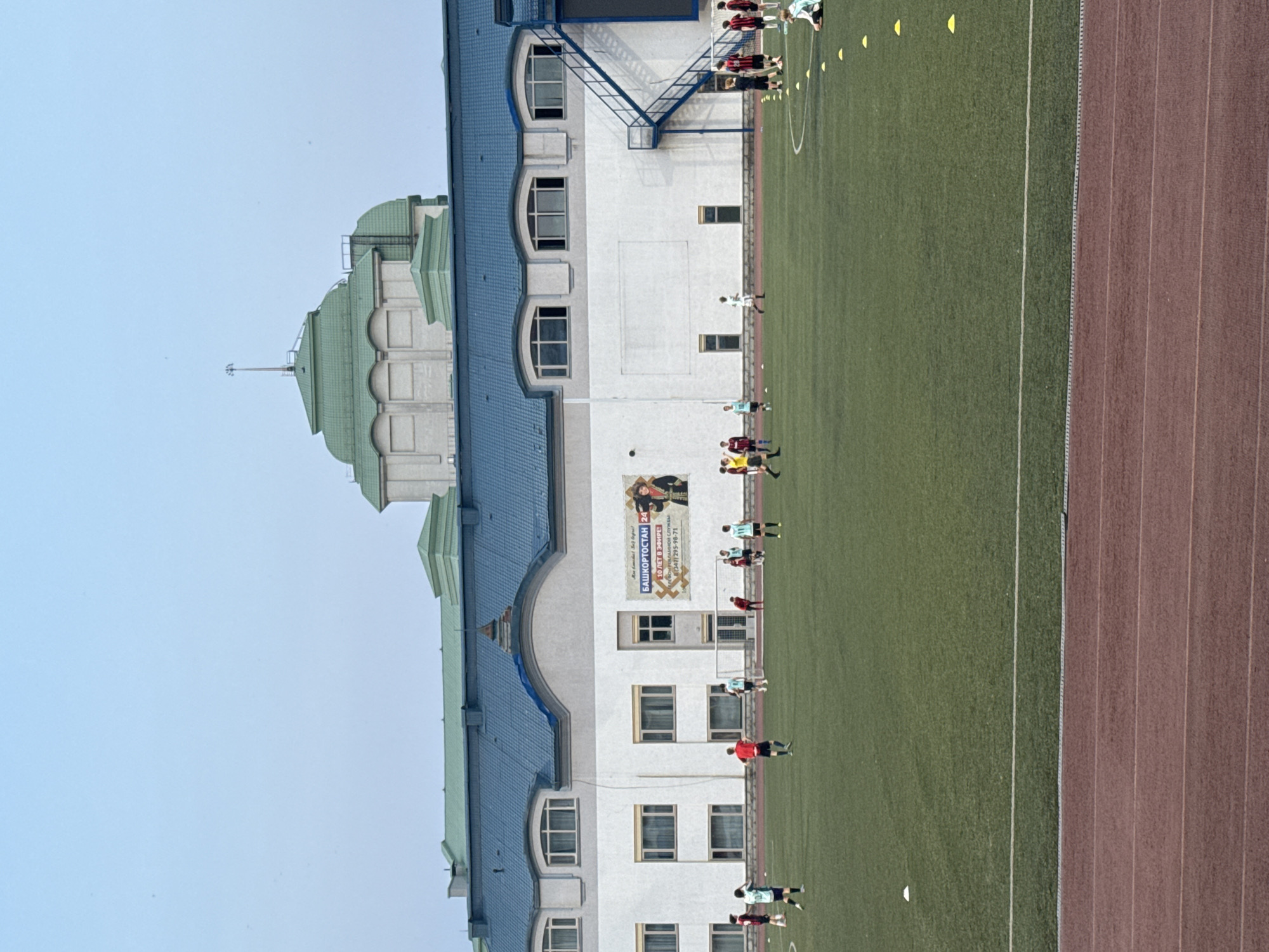 The image depicts an outdoor football (soccer) field at the Salavat Yulaev Sports Palace in Ufa, Russia. Several young males, dressed in football attire, are scattered across the green turf, engaging in what appears to be a training session. Some are running, while others stand in formation or are in the process of kicking a ball. A few individuals are seated on the field, observing the activity. The field is marked with white lines and dotted with yellow cones, indicating drills or practice zones. In the background, a large, modern building with a blue roof dominates the scene. This structure, part of the sports complex, features multiple windows and an external staircase. Atop the building, a distinctive green, multi-tiered dome can be seen, a prominent architectural feature. A large banner is displayed on the building's facade, featuring text and an image. The sky above is clear and pale blue, suggesting it is either early morning or late afternoon. The overall impression is of a sports facility actively in use for youth athletic training.