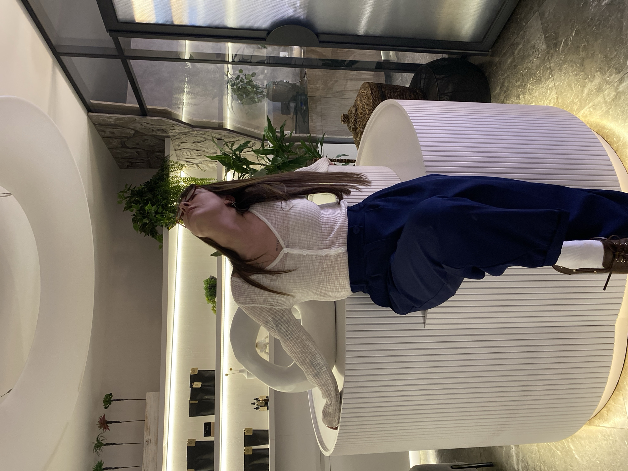 A full-length, vertically oriented photograph captures a young woman reposing in a modern, well-lit interior, likely a commercial or public space in Votkinsk, Russia. The image is rotated 90 degrees clockwise.
The woman, with long dark hair, wears a white, sheer, long-sleeved button-up top, dark blue wide-leg trousers, white socks, and brown laced shoes. Her head is tilted far back, and she gazes upwards, her mouth slightly open, suggesting a pose of relaxation or contemplation. A faint tattoo with text, possibly "mirror," is visible on her neck.
She is situated on a distinctive, curved white bench or structure featuring horizontal ridged patterns. To her left, another smooth white, arched architectural element is visible. The background features various elements suggesting a stylish interior design. There are green potted plants, some small and artificial, others larger, adding a touch of nature. Behind the woman, a display shelf with integrated lighting holds several small black pouches or boxes. Further back, a dark-framed glass partition or window with reflections is seen, along with sections of textured stone walling. A large, dark woven basket is also discernible in the upper right. The overall atmosphere is clean, contemporary, and well-designed, possibly a showroom or a modern cafe.
