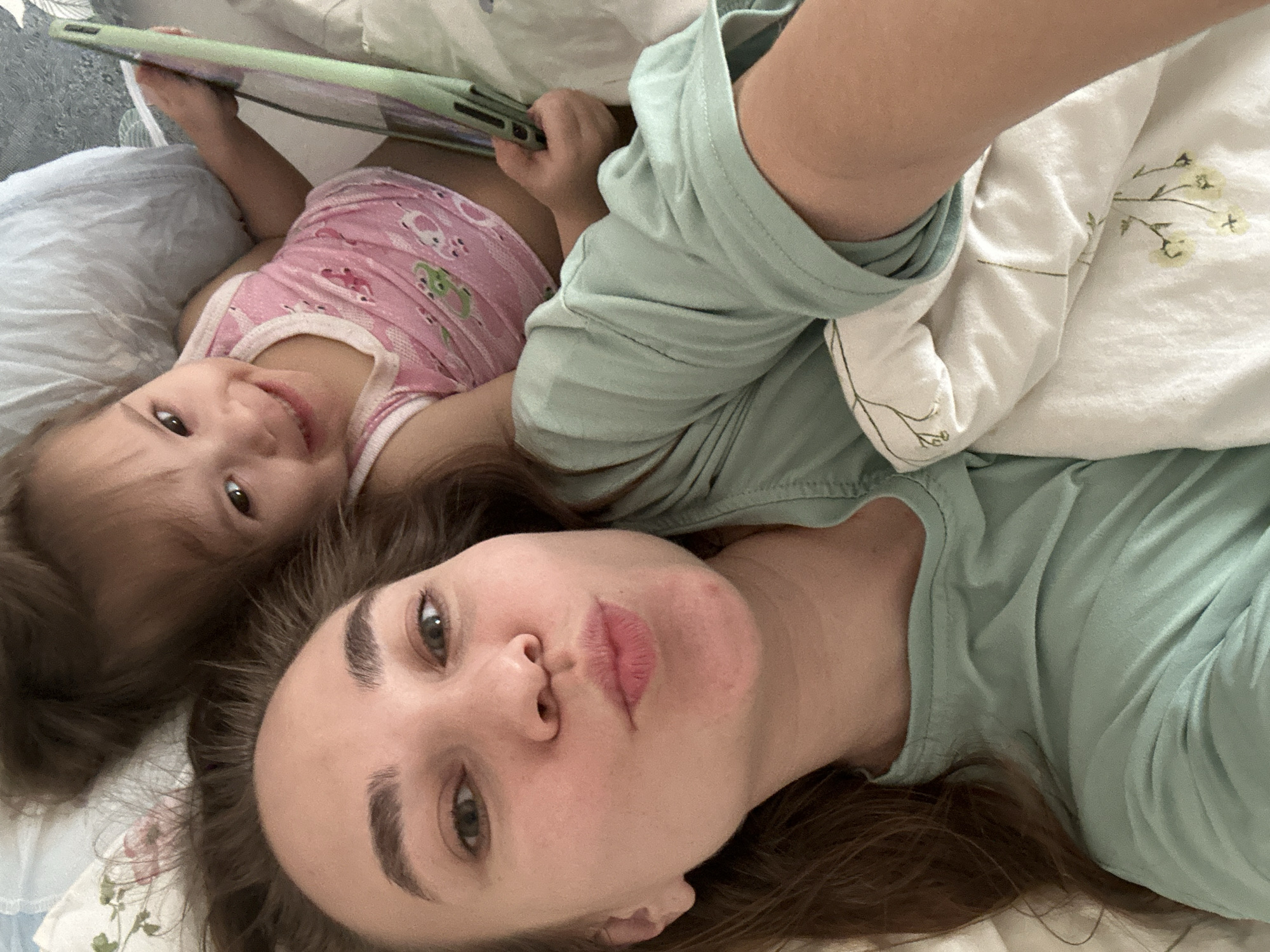 An overhead selfie captures an adult woman and a young child lying down together indoors, likely on a bed or sofa. The image is taken from above, with the subjects oriented diagonally across the frame.
The woman is in the foreground, lying on her back, looking directly at the camera with a playful expression, her lips slightly pursed in a "kissing" pose. She has long, dark brown hair spread out around her head and is wearing a light green long-sleeved top. Her skin appears fair, and she has well-defined eyebrows.
Behind and to the left of the woman is a young child, appearing to be a toddler or preschooler. The child is also lying down, looking towards the camera with a joyful smile. They have dark brown hair and are wearing a pink tank top adorned with repeating white trim and cartoon animal patterns, possibly frogs or rabbits. The child's left hand is holding a light green-cased tablet or large smartphone, which is angled towards the top left of the frame.
The setting appears to be a cozy indoor space, with soft furnishings visible. A light blue or gray pillow is under the child's head, and parts of a white blanket with delicate green floral patterns are visible towards the upper right. The lighting is soft and natural, suggesting daytime. The overall mood is intimate, relaxed, and affectionate. There is no visible text in the image.