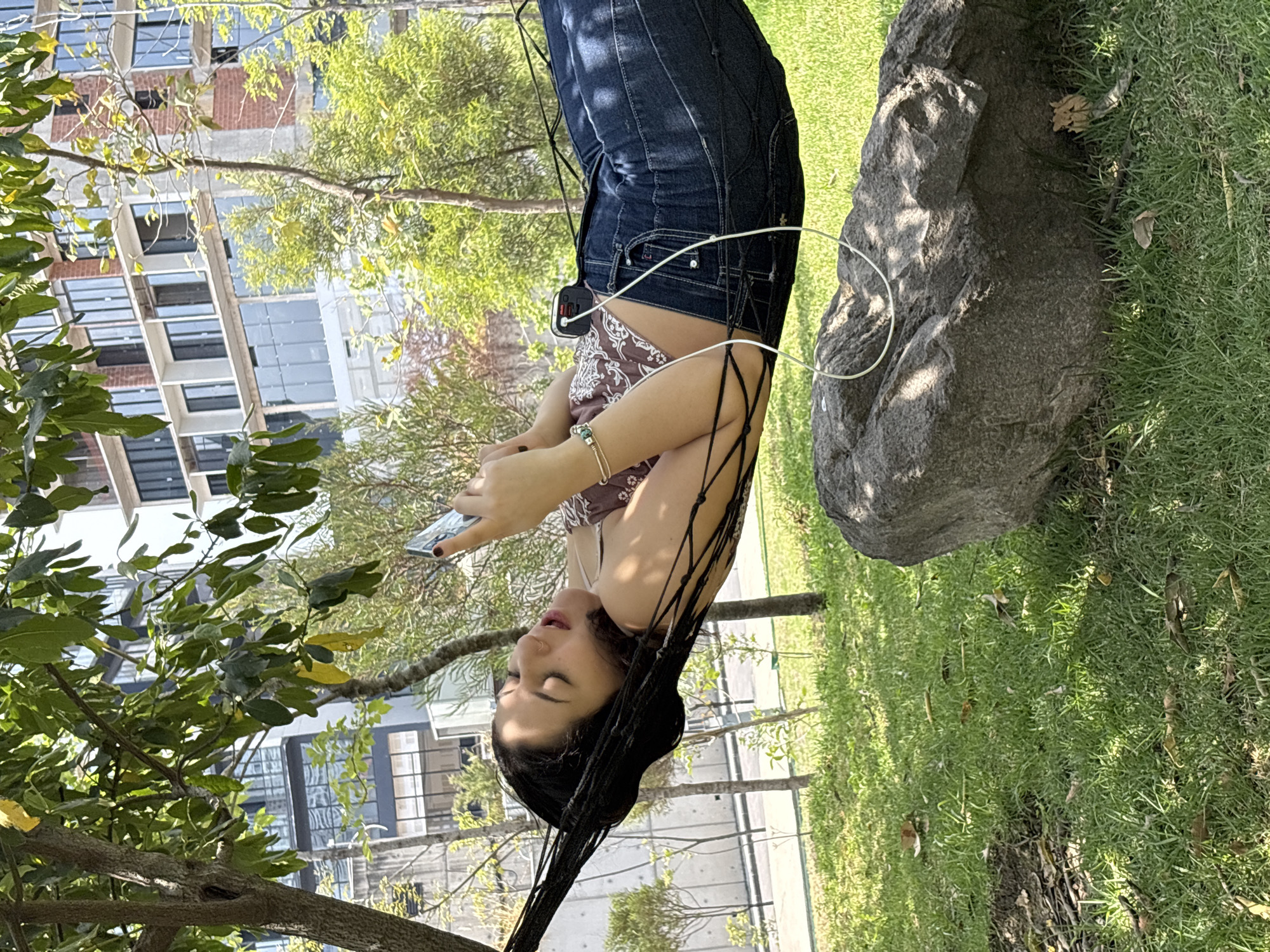 A woman in jeans and a brown top lies in a hammock between a tree and a large rock, looking at her phone. She is in a park-like setting, with green grass and trees visible in the background. Modern buildings are visible in the distance, suggesting an urban environment, likely in the city of Jesús del Monte, Mexico. A white cable connects to the phone.