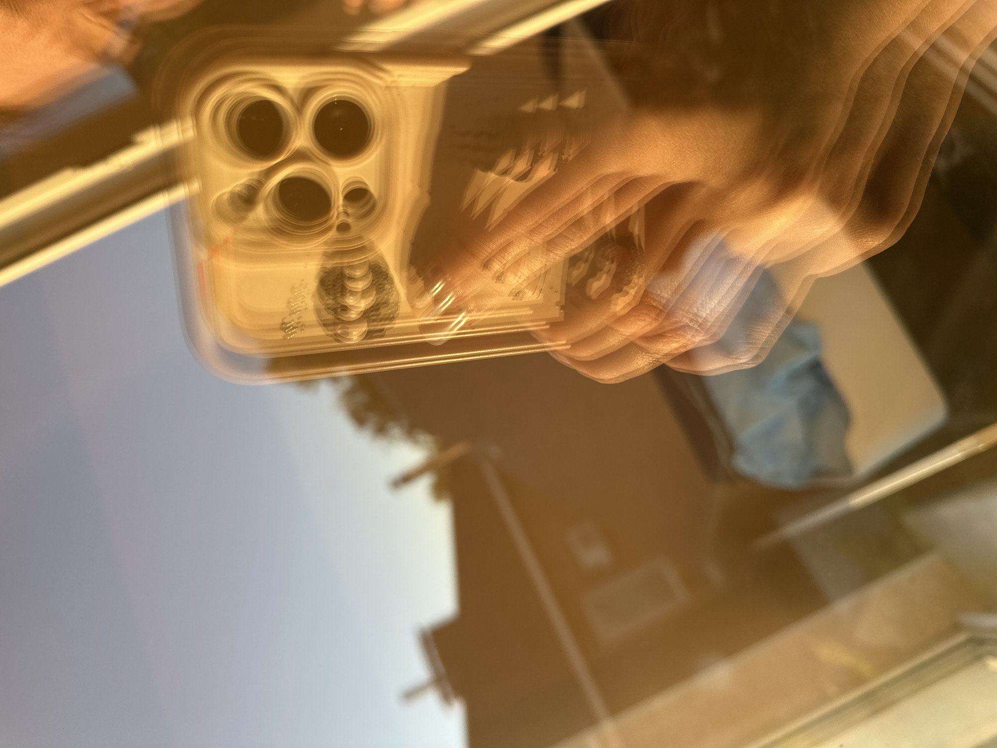 The image is a close-up, slightly blurred photograph taken from the perspective of someone holding a smartphone. The phone, which has a clear case and a distinctive camera array on the back, is the primary subject in the foreground, held by a hand with visible fingers and fingernails. The warm, golden light suggests it may be dusk or dawn, casting long shadows and reflections.
In the background, a blurred reflection shows the exterior of a building, possibly a house, with visible windows and architectural features. The sky above is a pale blue, transitioning to a warmer hue near the horizon, indicating it might be sunset or sunrise. The overall impression is that of a selfie taken through a window or a reflective surface, possibly in an urban or suburban environment. There is no information in the image to suggest the location is Nizhny Novgorod, Russia.