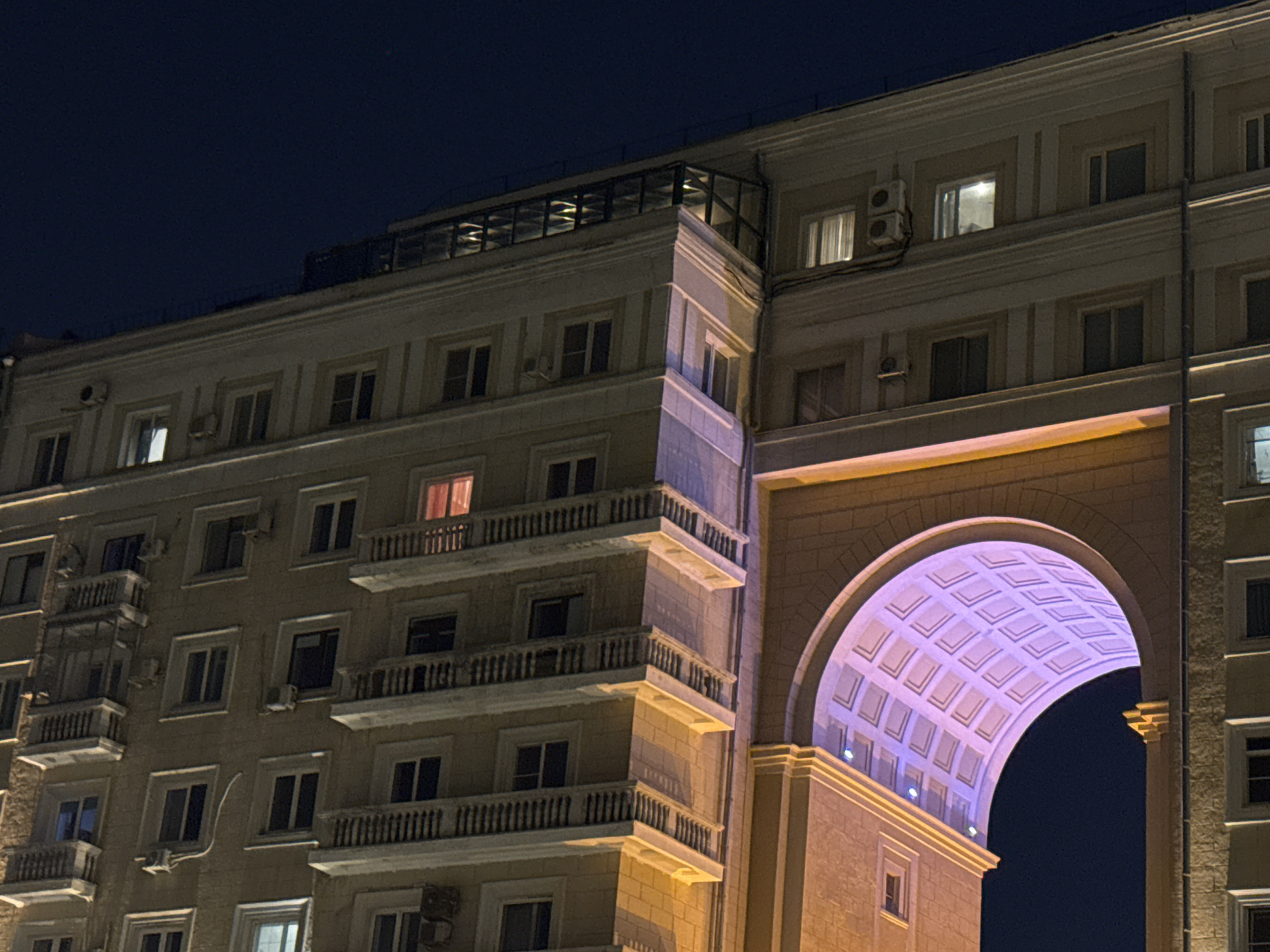 The image shows a multi-story building at night in Moscow, Russia. The building is beige with rows of windows, some with balconies and air conditioning units. On the right, a large arched opening is illuminated with a purple light, contrasting with the dark sky visible through the arch. The architecture is classical in style.