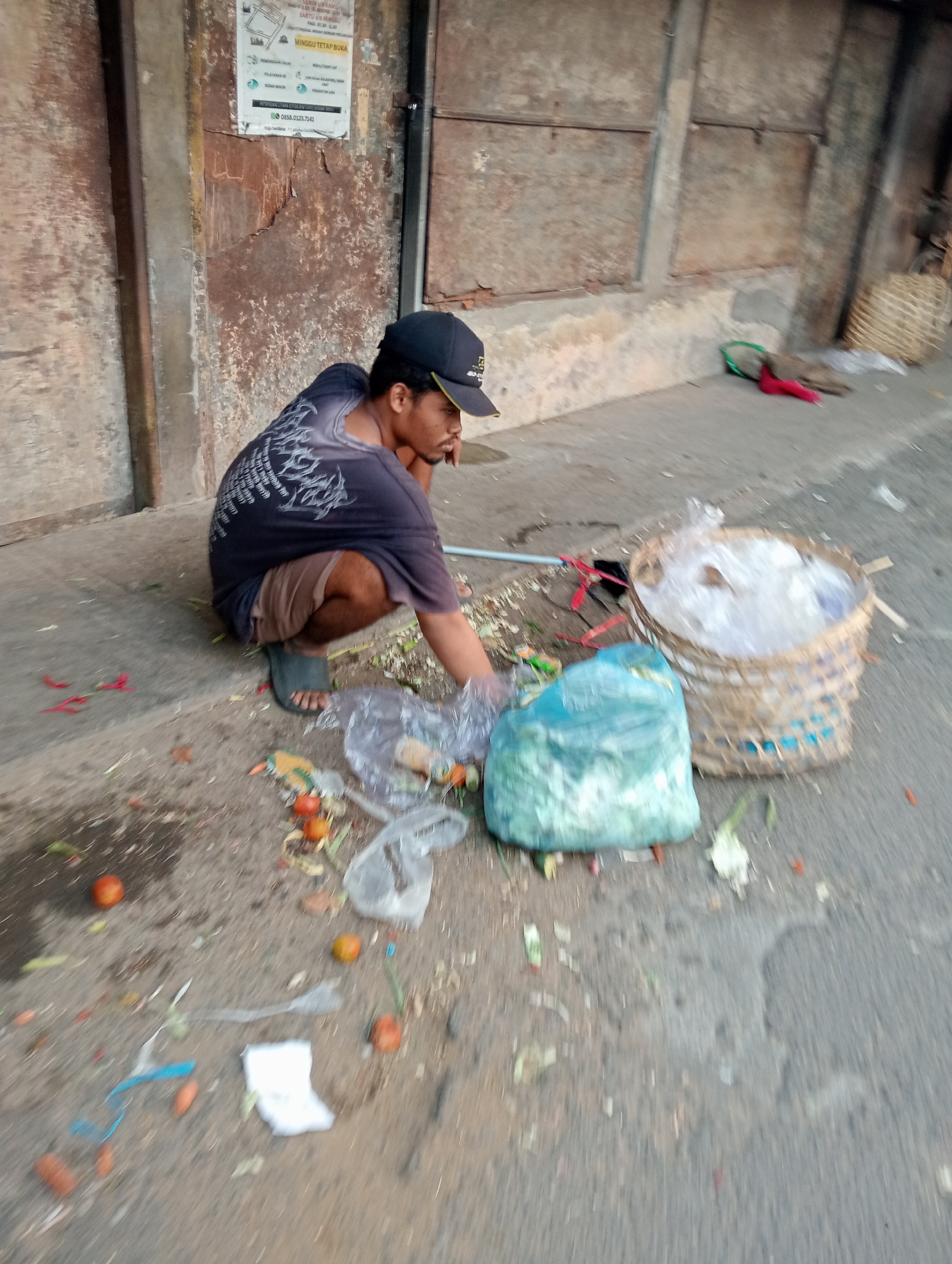 A man wearing a dark t-shirt, brown shorts, and a black baseball cap is squatting on a dirty sidewalk. He appears to be sorting through garbage that is scattered on the ground. In front of him are two bins: a woven basket filled with what appears to be plastic bags, and a large, light blue plastic bag. Debris, including vegetables, plastic, and red chili peppers, is strewn across the ground around him. In the background, there is a weathered, rusty wall and a concrete ledge. The scene suggests a public space in Tanubayan, Indonesia, possibly a market or roadside area, where waste management is minimal.