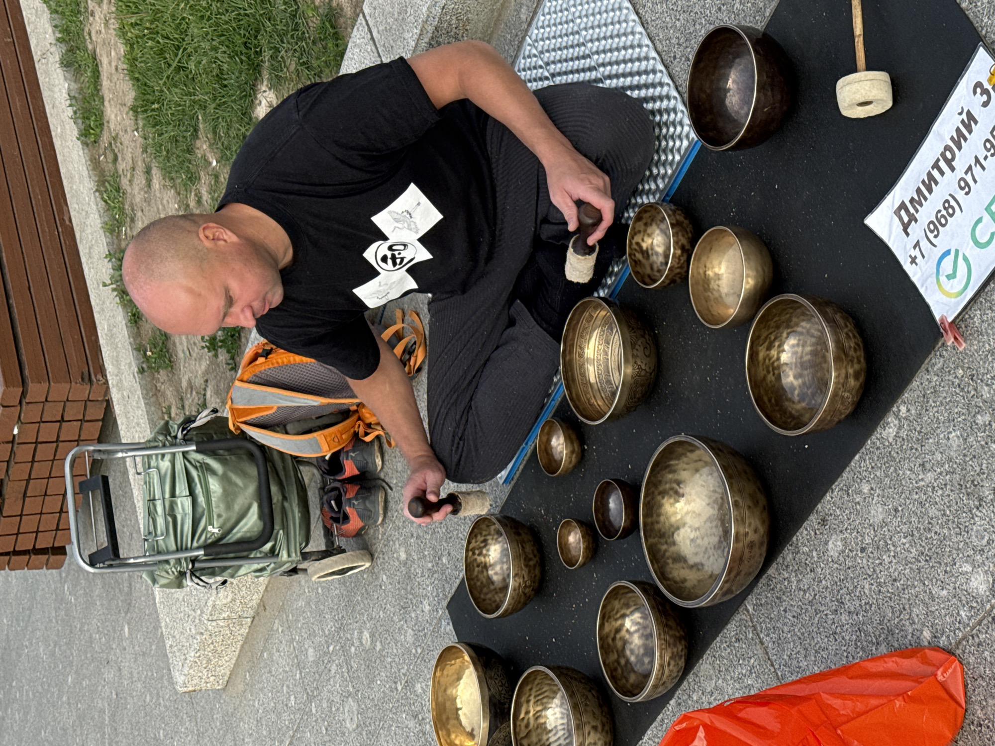 This overhead shot shows a bald man sitting cross-legged on a grey paved surface, demonstrating or playing a collection of singing bowls. He is wearing a black t-shirt with a white graphic print and dark trousers. In his right hand, he holds a mallet, poised over one of the many brass singing bowls arranged on a black mat in front of him.
The mat displays at least ten singing bowls of various sizes, along with additional mallets. To the man's left, an olive green backpack is secured to a metal trolley, and a pair of dark sneakers with orange accents rests on the ground. The immediate surroundings include a strip of grass and part of a wooden bench, indicating an outdoor, public setting.
On the black mat, a white sign with Russian text is visible. It reads: "Дмитрий З" (Dmitriy Z), followed by a partial phone number "+7 (968) 971-9", and the letters "СБ" with a green checkmark logo, possibly indicating a payment method or contact detail. The scene appears to be taking place during the daytime under clear weather conditions. The man has a focused expression, engaging with the musical instruments. Given the context, this event likely occurs in Moscow, Russia.