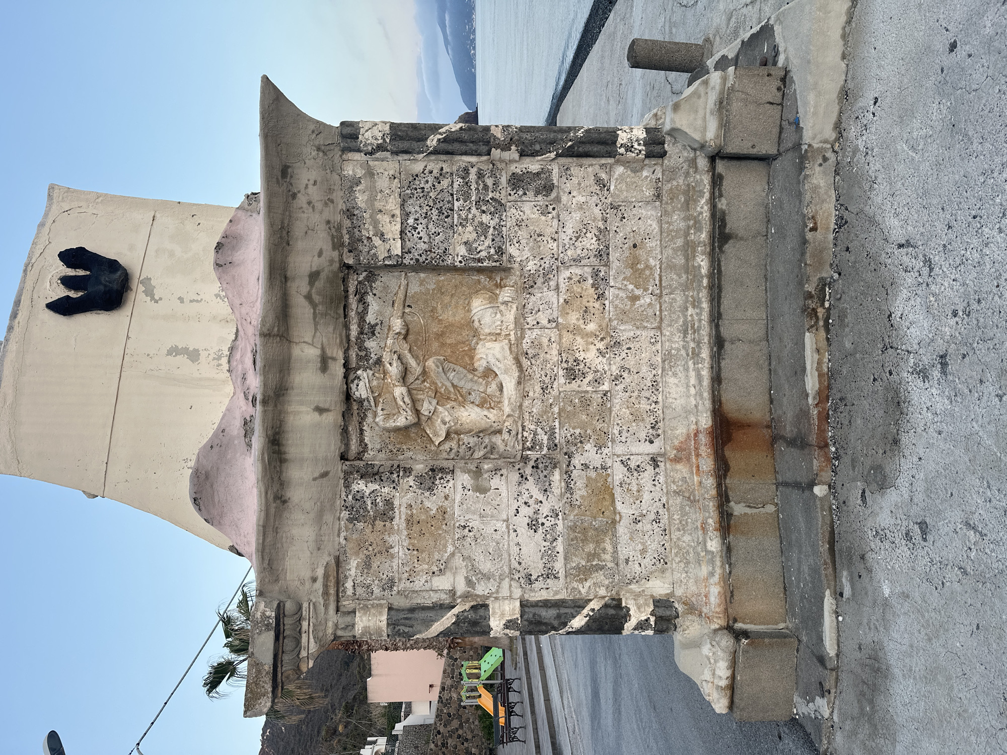 This media file presents a weathered stone monument or building facade set against a clear blue sky and a coastal landscape. The central feature is a substantial, rectangular stone structure, showing considerable age and wear, with a rough, textured surface displaying various shades of grey, beige, and reddish-brown stains. Embedded within this structure is a detailed bas-relief carving.
The carving depicts two stylized human-like figures in a dynamic interaction. The figure on the right appears to be seated or kneeling, wearing a distinct headdress. The other figure, positioned to the left, seems to be in a more active stance, possibly holding a spear or staff. The carving, though showing signs of erosion, retains its intricate details.
Above this stone structure, a lighter-colored wall, possibly stuccoed and painted pale beige with a faded pink patch, rises towards the sky. Affixed to this wall is a striking, black, abstract sculpture resembling a large claw, a wing, or a stylized letter 'E'. The top edge of the structure features a pointed, fin-like architectural element.
In the upper background, beyond the structure, a calm, expansive body of water, likely the sea, stretches towards a distant landmass or island with a rugged coastline, all under a bright, clear blue sky. In the lower background, to the left, residential buildings and dark, rocky hills are visible. Closer to the viewer, a vibrant children's playground with green slides, yellow platforms, and dark benches is situated next to a paved road. A few palm trees are also visible.
The overall scene conveys a sense of history and endurance, with the heavily weathered stone indicating significant age and exposure to the elements. The bright blue sky suggests it is a sunny day. No visible text is present in the image. The combination of ancient-looking stone, a vibrant playground, and a coastal setting hints at a location like Lipari, Italy, where history and modern life intertwine by the sea.