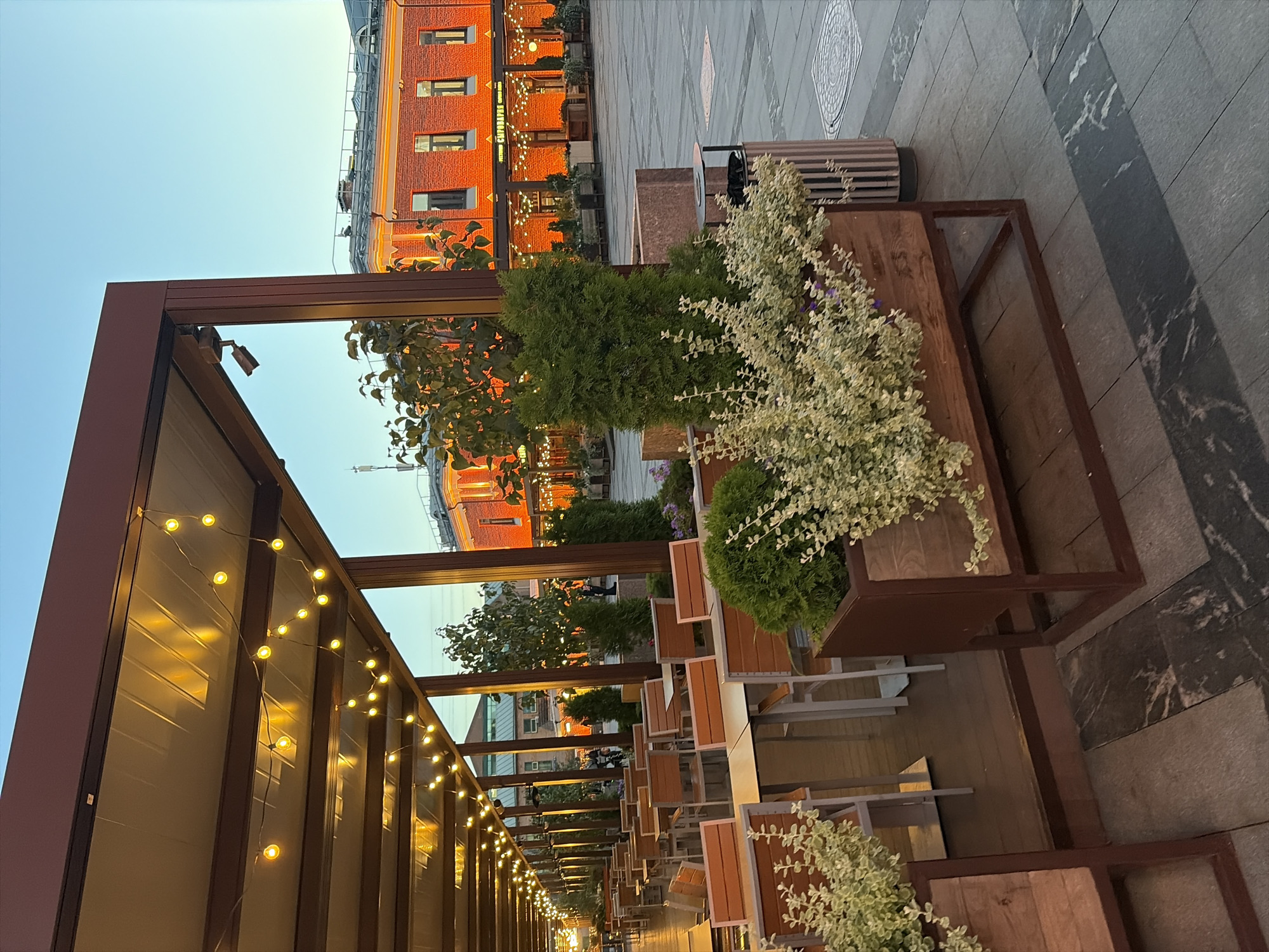 This image depicts an outdoor seating area of a restaurant or cafe in Moscow, Russia, during what appears to be dusk. The area is covered by a large, modern pergola with a metallic roof supported by brown wooden beams. String lights with warm yellow bulbs are strung along the underside of the pergola, illuminating the seating area.
The seating consists of numerous tables with light-colored tabletops and chairs with wooden slats and metal frames. The tables and chairs are arranged in rows, extending into the distance. Several planters containing lush green bushes and smaller plants with light-colored leaves are strategically placed throughout the seating area.
In the background, a red brick building with several windows is visible. The building is illuminated with warm orange light, suggesting interior lighting or external decorative lighting. The architecture of the building appears to be historic, with multiple stories and a visible rooftop. The ground surface is paved with light-colored square tiles, and there are darker rectangular stone tiles forming a pattern.
The sky above is a clear, pale blue, characteristic of twilight. The overall scene conveys a sense of a relaxed, ambient outdoor dining experience. A few indistinct figures of people can be seen in the distance, suggesting the establishment is either open or preparing to open for the evening. A trash receptacle with a striped pattern is positioned near one of the planters. The image is captured from a slightly low angle, looking across the seating area towards the building.