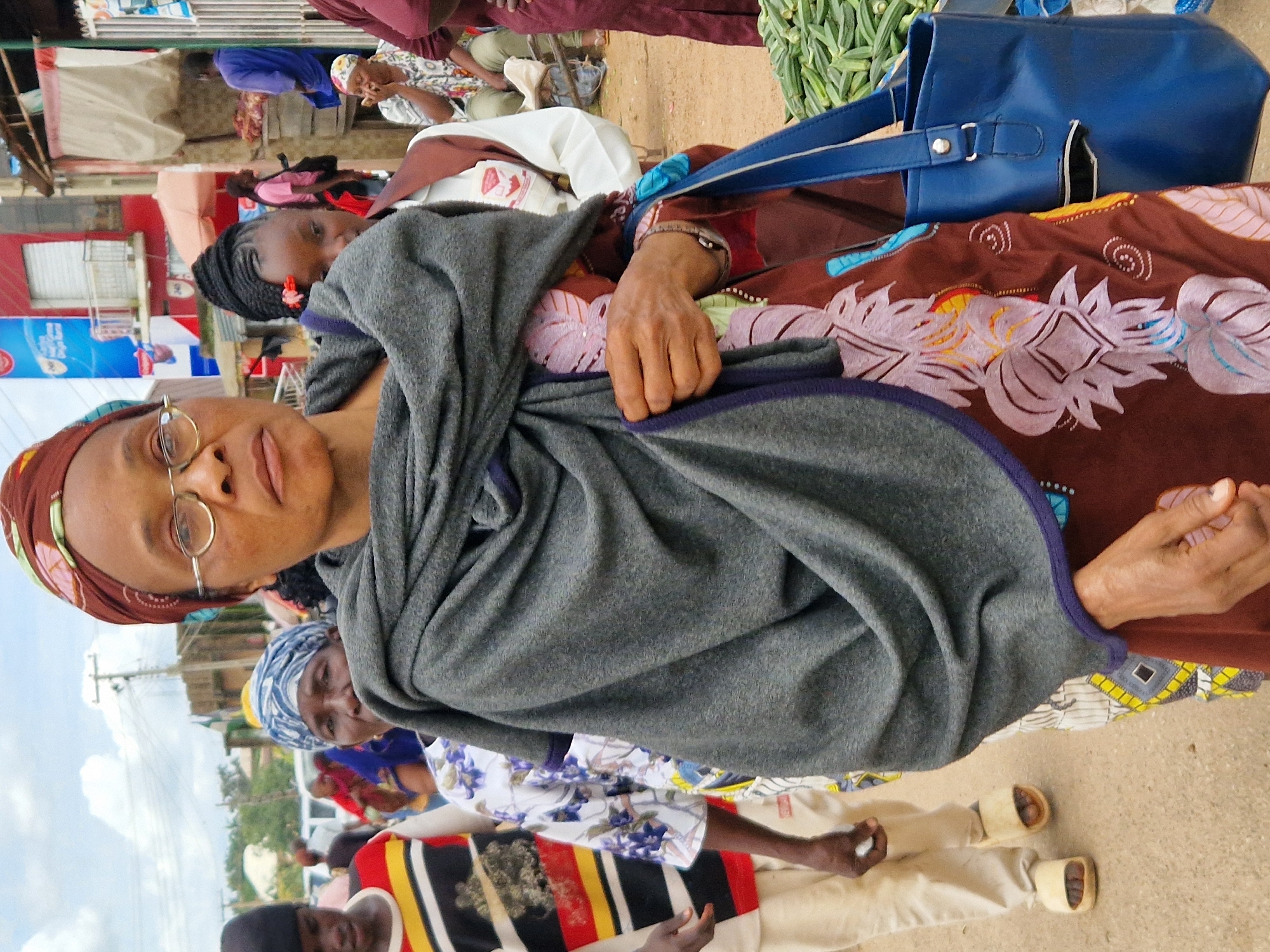 In a Nigerian street scene in Kabong, Nigeria, a woman with a patterned headscarf and glasses is the primary subject. She is wrapped in a large, grey shawl, obscuring most of her clothing, but a glimpse of a brown garment with floral patterns is visible. A blue handbag is slung over her right shoulder. To her left, a person wearing a striped shirt with red, yellow, black, and white bands is partially visible, as is another person in the background wearing a blue and white headscarf. Further back, several other individuals are present, some walking, some standing, and some seated. In the background, there are market stalls, buildings with red and white facades, and a blue advertisement banner. The ground appears to be dirt or gravel, and a pile of okra is visible to the right of the frame. The sky is overcast with white clouds.